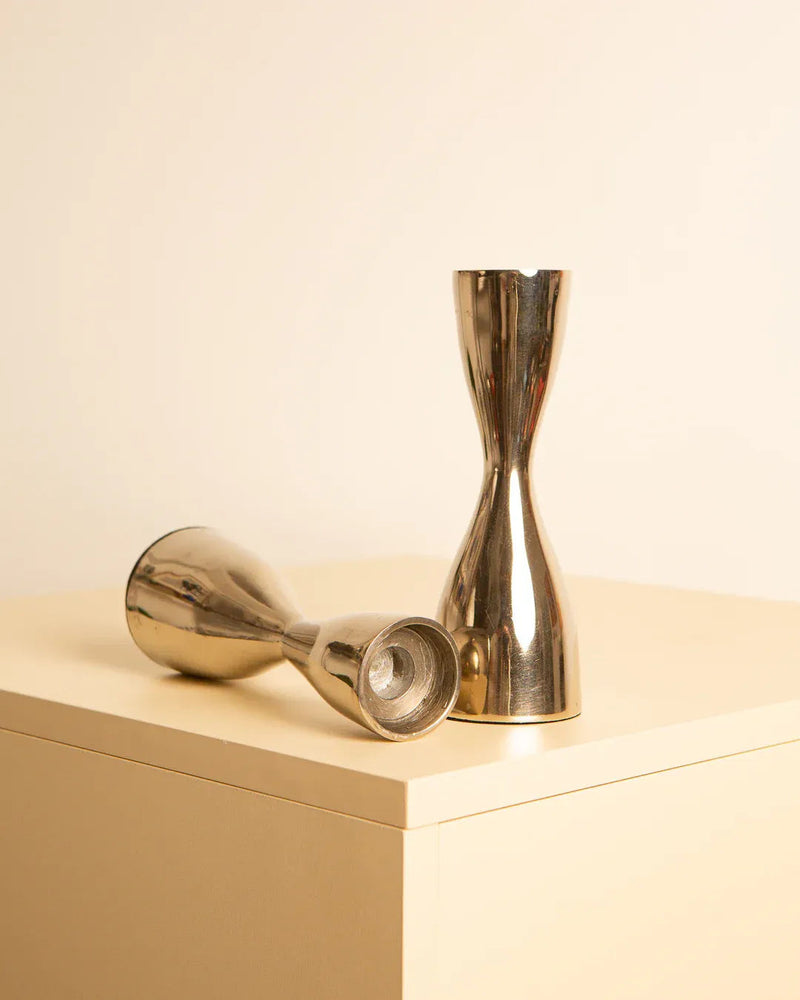 Two metallic hourglass-shaped objects, reminiscent of 1980s Italy, are displayed on a beige surface against a cream background. One object stands upright while the other lies horizontally beside it. The objects have a reflective, shiny finish, resembling elegant metal candleholders. These are the Pair of “Hourglass” candleholders in Italian metal 80's by Treaptyque.