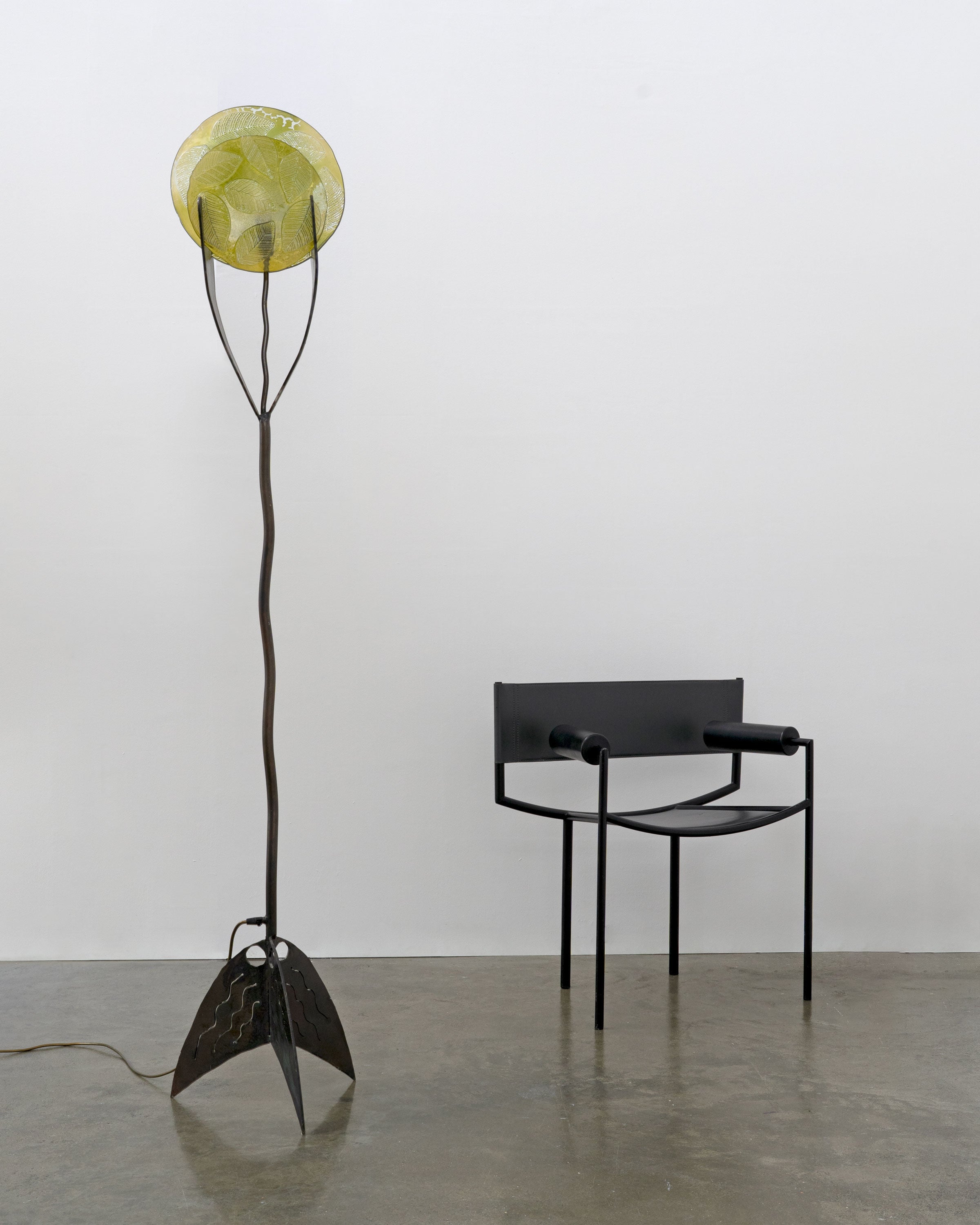 A black metal chair with cylindrical armrests sits beside a Tom Bogle Brutalist Wrought Iron and Cast Glass Lamp (Norway, c.1970), featuring a yellow circular shade and wavy tripod base, on a polished concrete floor against a plain white wall.