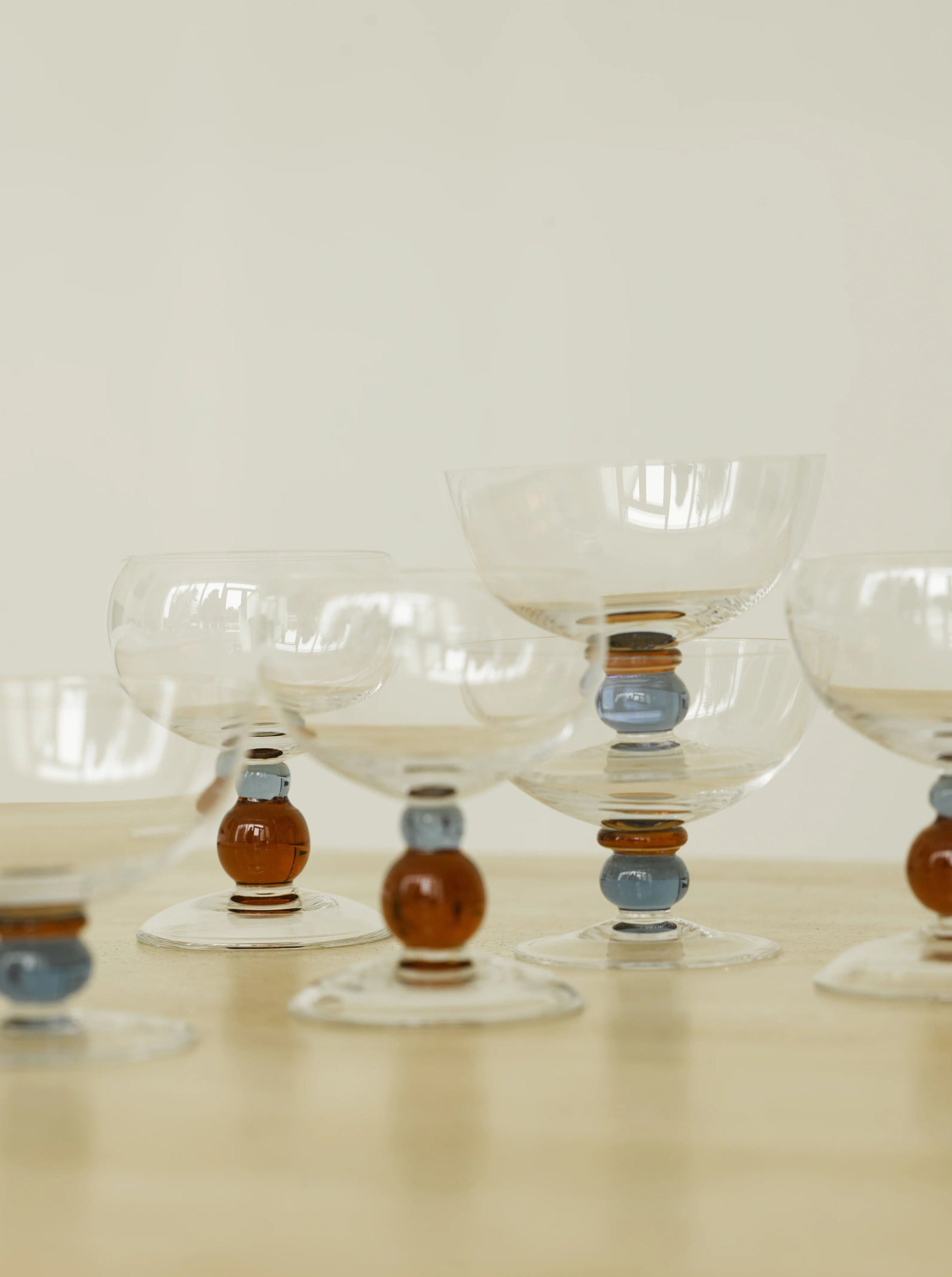 A set of four clear glass goblets with decorative stems, featuring brown and blue spherical accents, arranged in a staggered formation on a light wooden surface against a plain background. These handmade Noam Wine Glasses by Akua Objects have a classic, elegant design perfect for festive seasons.