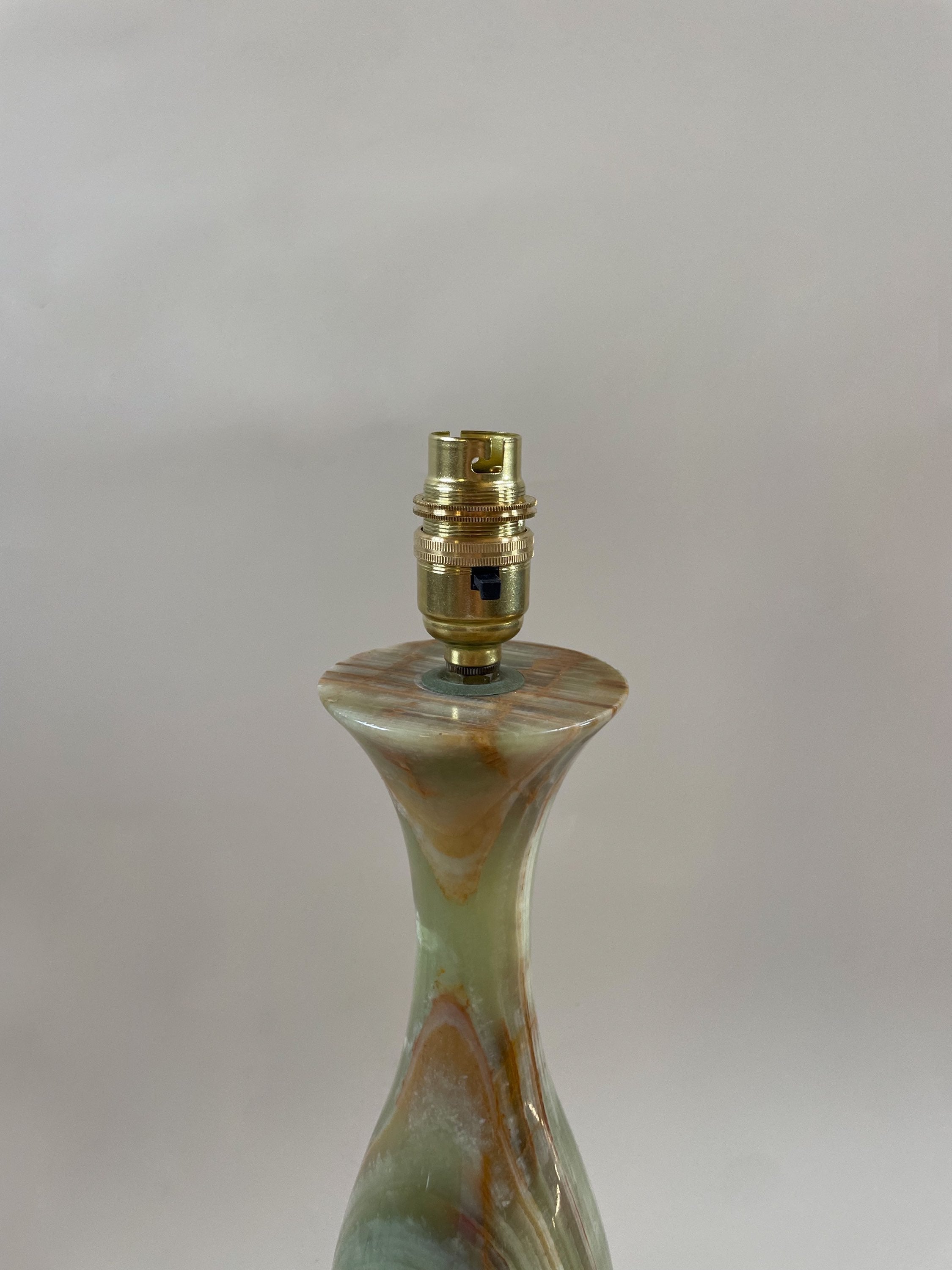 A vintage Maud Vaughan Green Onyx Lamp C.1970 tapers upward to a golden bulb socket with subtle brown, green, and beige swirls in the base. The image captures the top part of this exquisite piece against a plain, light gray background.