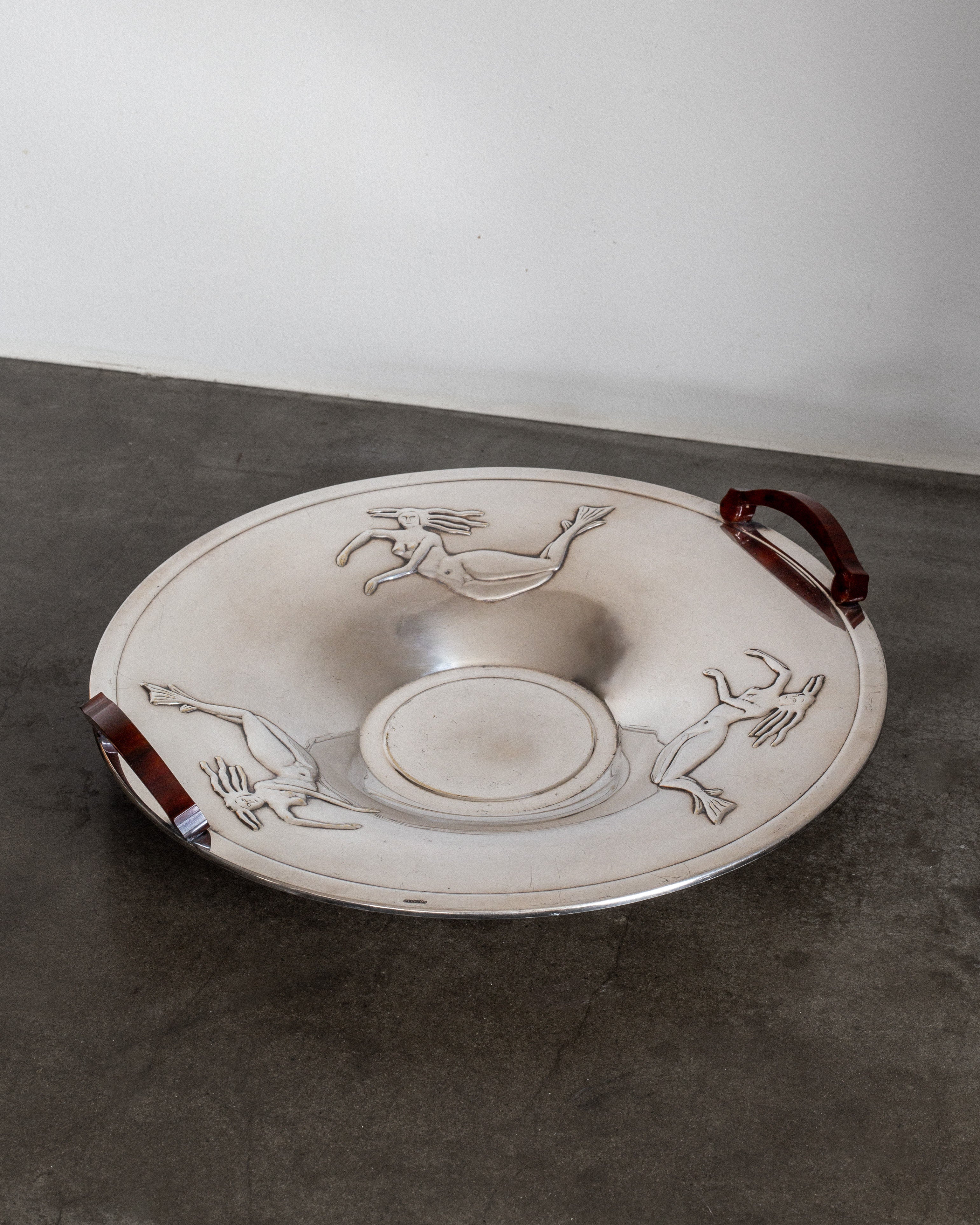 A large, shallow silver-plated dish from the 1930s by Spigel, featuring two Bakelite handles and three engraved animals around the rim, is displayed on a dark stone floor against a white wall.