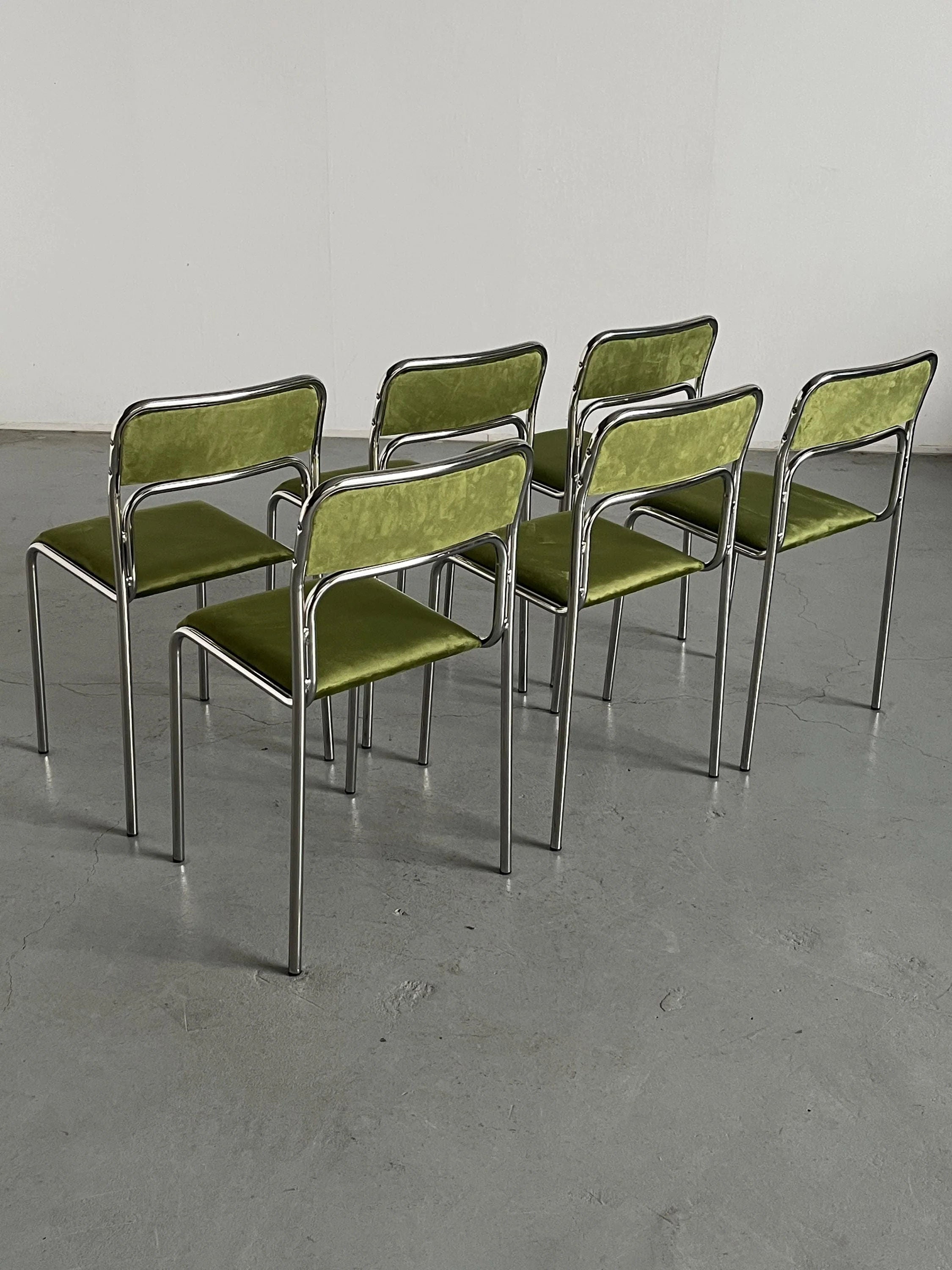 The Cherry Cargo set of 6 Bauhaus Chrome and Green Velvet Chairs are neatly arranged in two rows of three on a gray concrete floor against a plain white wall, showcasing their modern, minimalist design.