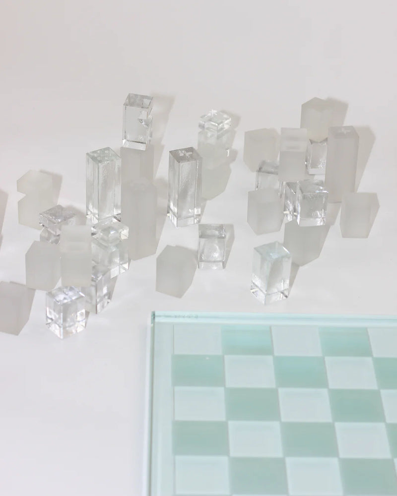 The Boga Avante Shop's Crystal Chess Set features a transparent glass board with clear and frosted, rectangular prism-shaped pieces elegantly displayed off the board on a pristine white surface, exuding minimalist sophistication.