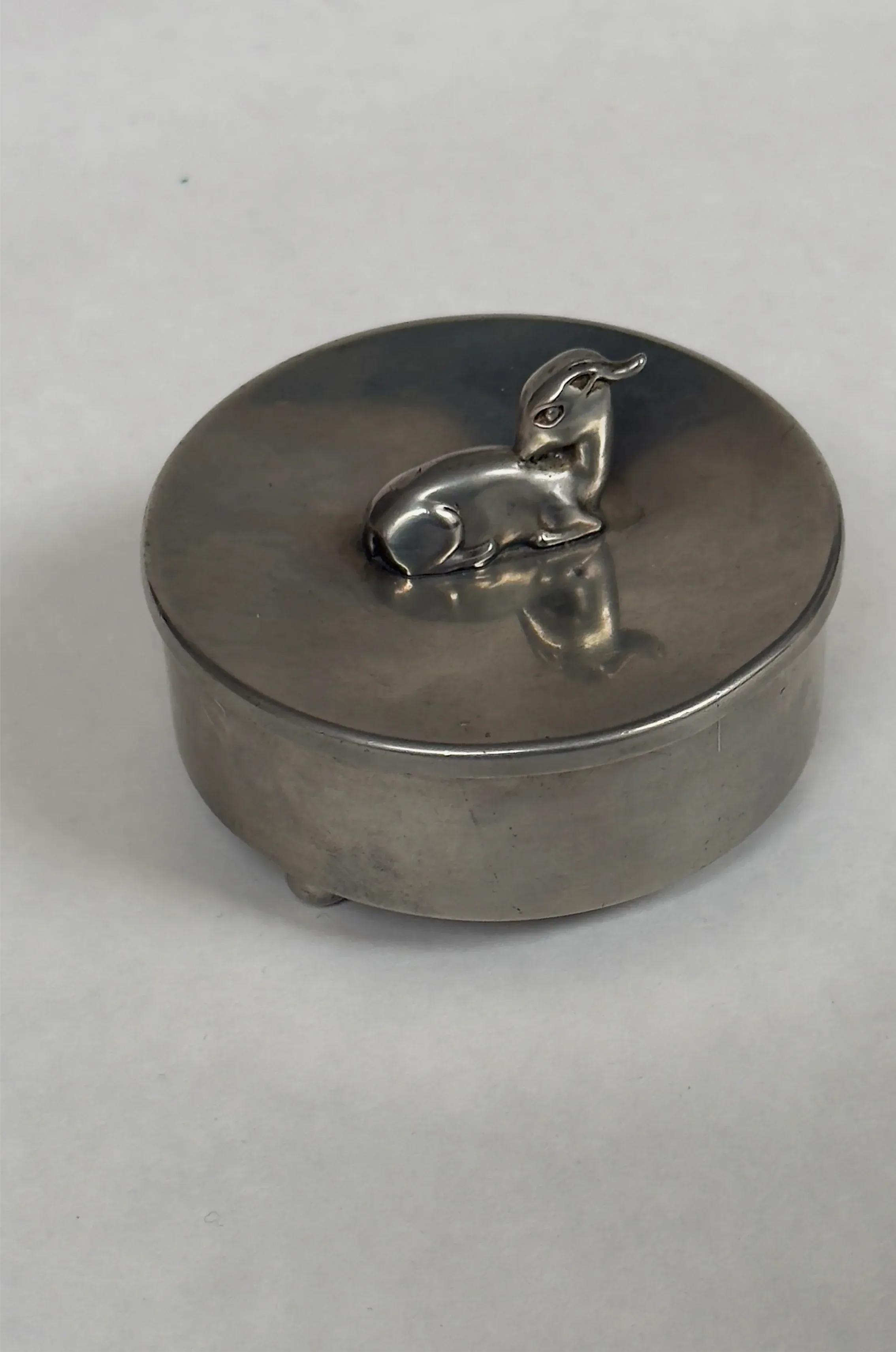 The Galerie Storm Vintage Pewter Trinket Box is a round, silver metal container with a sculpted rabbit figure on the lid, casting a beautiful silhouette on its reflective surface. Ideal for storing delicate jewelry.