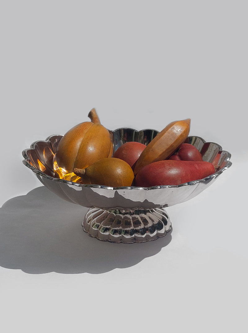 The Large Fruit Basket from Les Objoies showcases a variety of small squash and gourd-shaped objects in shades of brown, orange, and red. With its elegant design and reflective fluted edges, the centerpiece stands out against a plain gray background. The scene is minimalistic and well-lit.