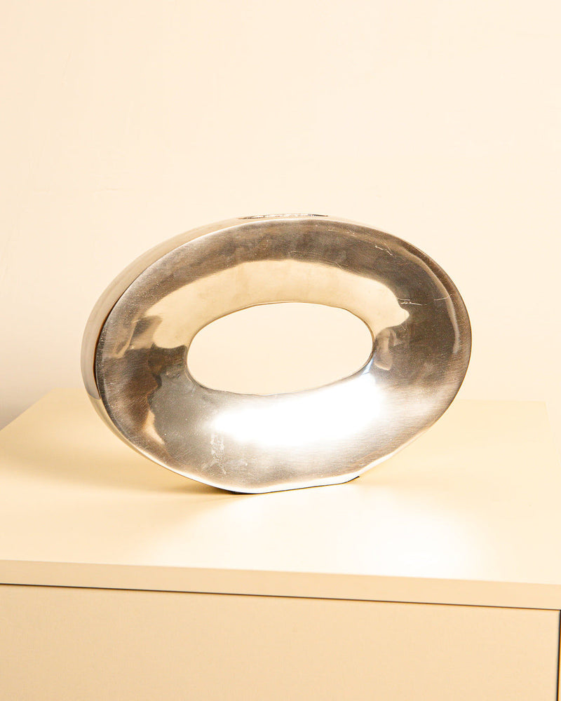 The "Ovale" Aluminum 80's sculpture by Treaptyque, a shiny oval piece with a large hollow center, sits on a cream surface against a plain light background, evoking vintage aluminum vase design.