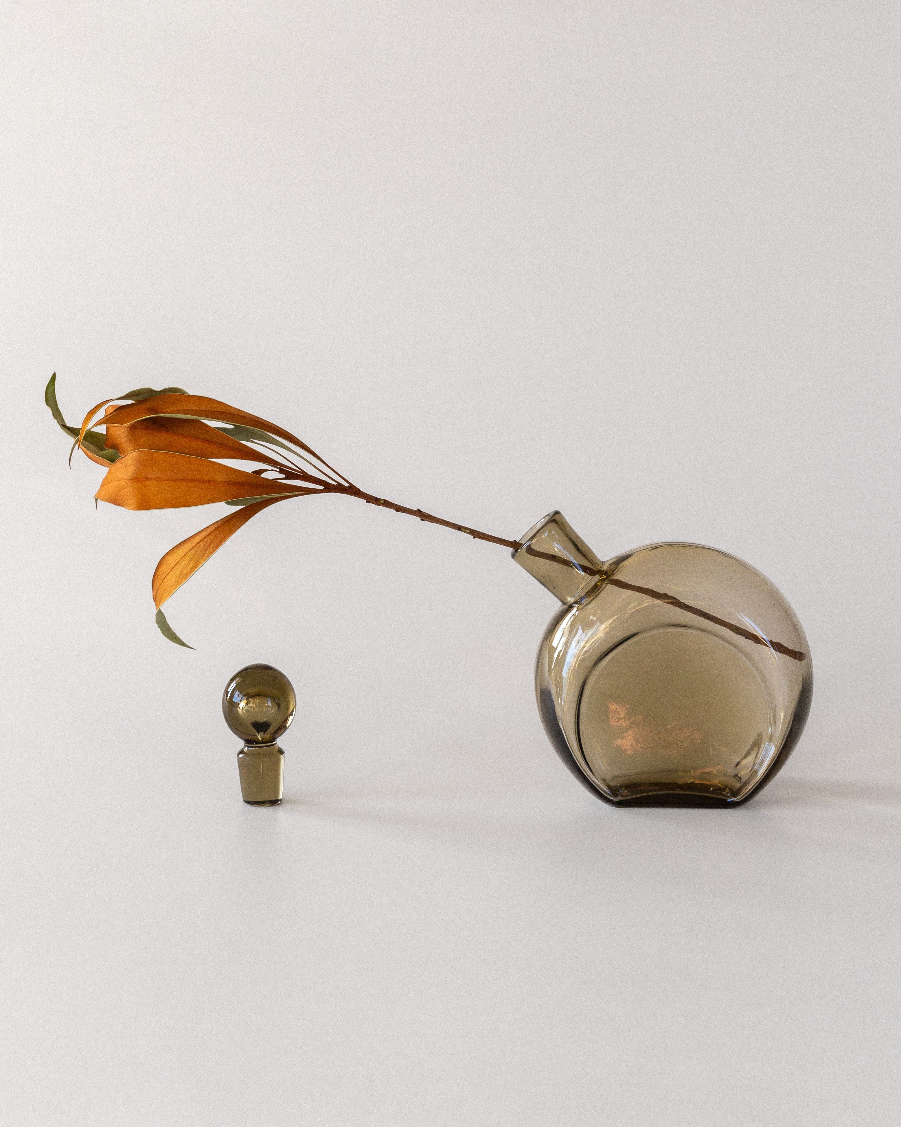 The Spigel Glass Decanter by Harald Notini from the 1930s, with a transparent round shape and narrow neck, holds a smoky brown leafy branch at an angle, embodying Swedish modernism. A small spherical glass object sits beside it on a cylindrical base against a soft gray background.