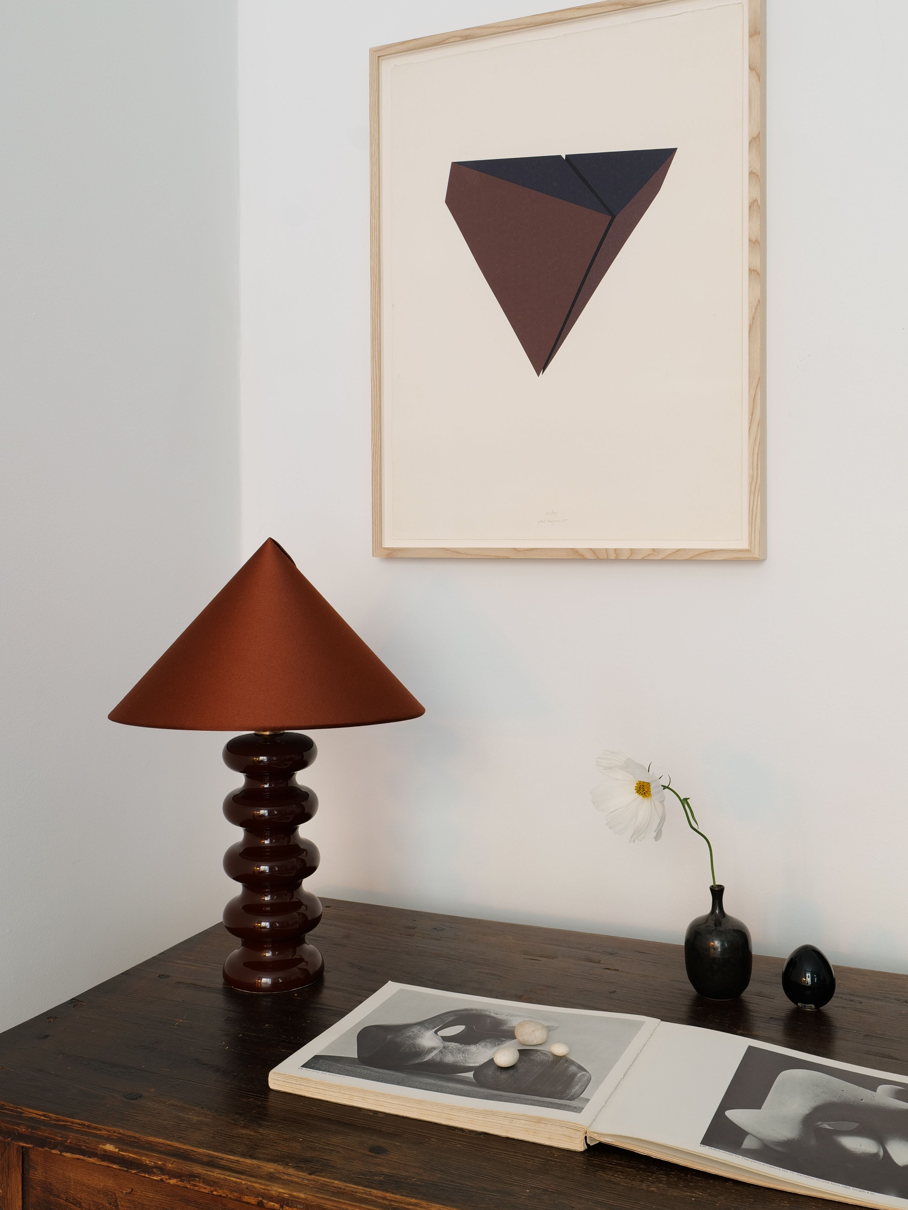 A cozy corner displays a Sculptural Ceramic Lamp by Collection apart with a handmade lampshade on a wooden table. Above, a framed abstract art piece with triangular shapes hangs on the wall. The table holds an open book with black-and-white photos, a single daisy in a vase, and two small stones.