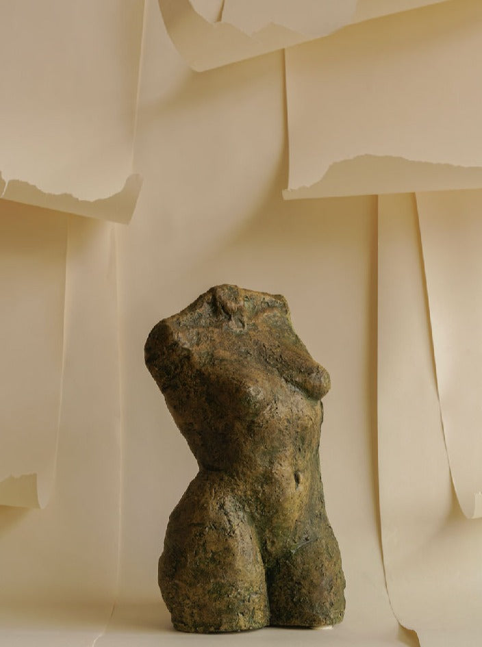 A Women Torso Sculpture of the Les Objets Amsterdam brand is centered in front of a backdrop of layered, slightly torn beige sheets of paper. The sculpture and wrinkled papers create a textured, artistic composition with a muted color palette.