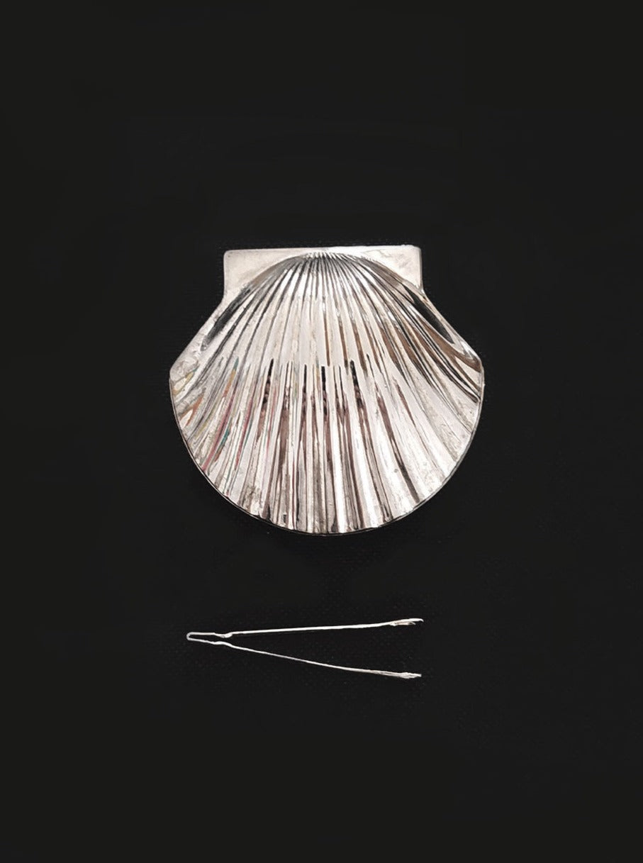 A large, shiny metal brooch shaped like a scallop shell, positioned above a Les Objoies Small Shell Box, against a plain black background.