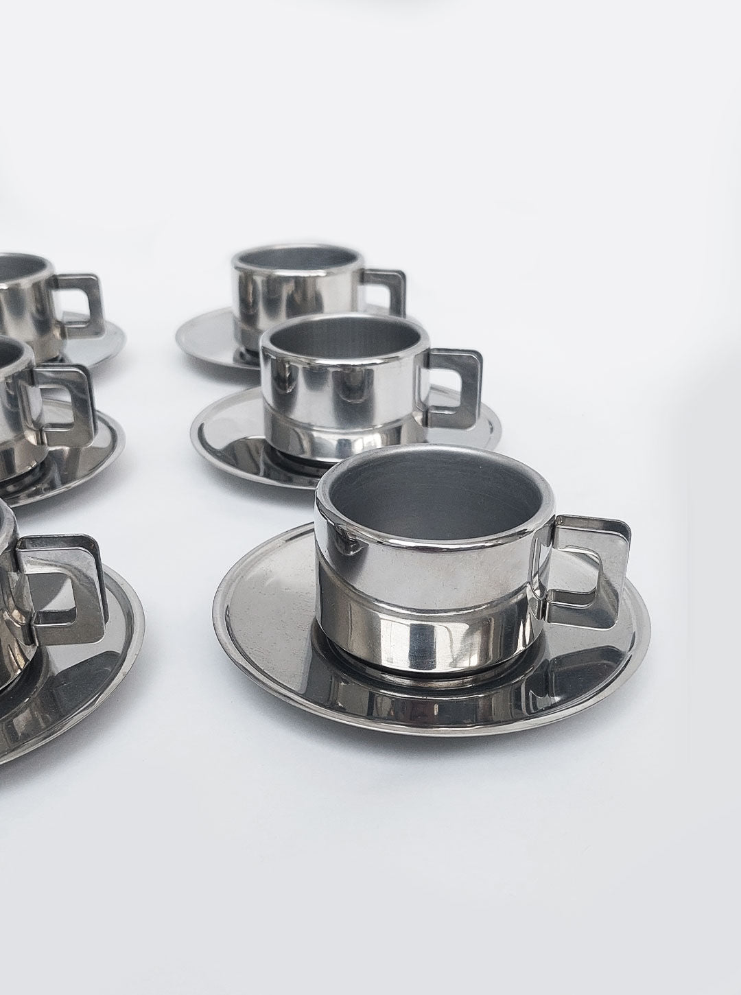 A Les Objoies Set of 6 Espresso Cups with matching saucers arranged in rows on a white surface. The cups have a shiny finish and rectangular handles.