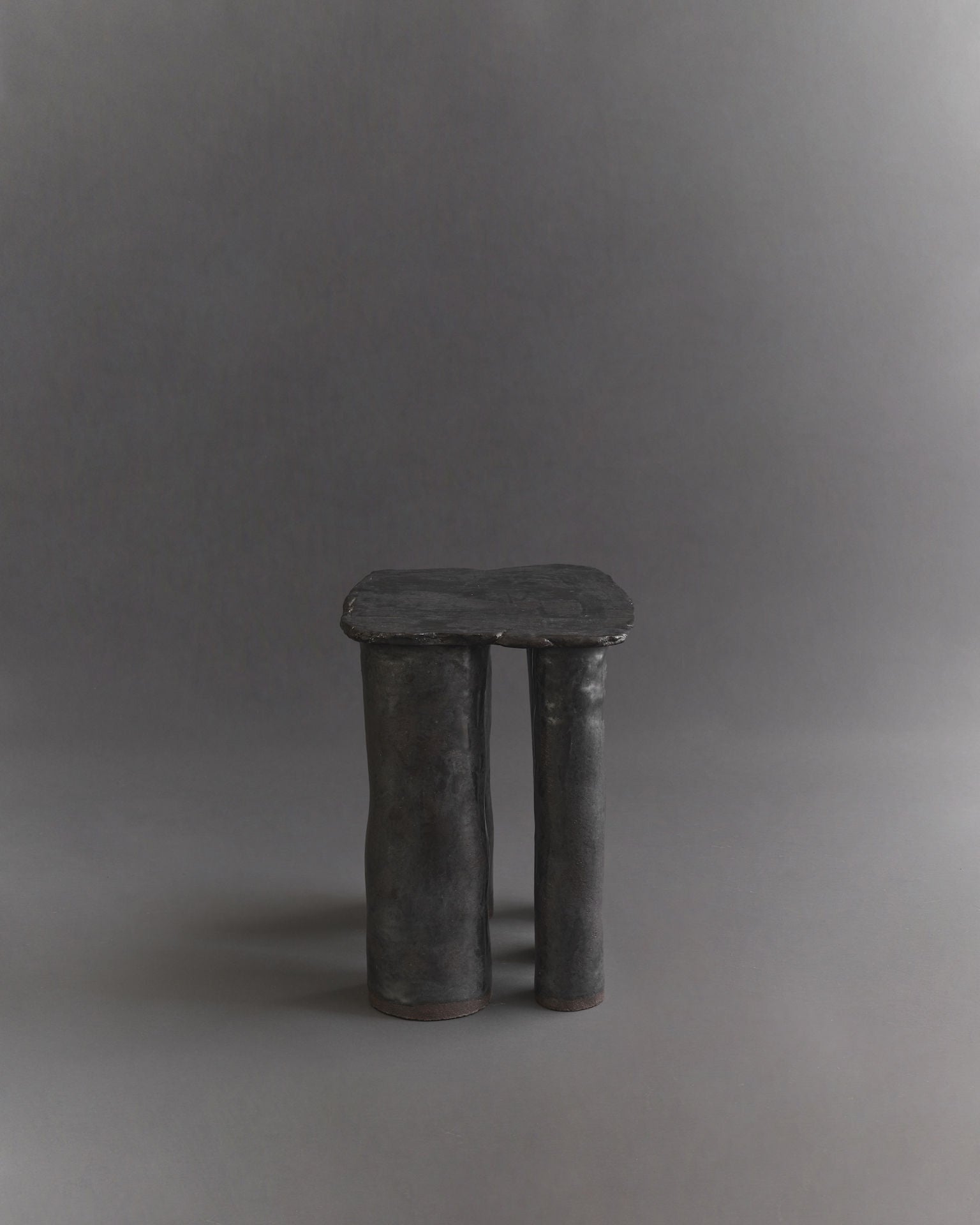 The Cleo Ceramic Side Table by Ombia Studio is a small, artistic black table with a rectangular top and textured glaze, supported by two thick cylindrical legs. This handmade piece stands out against the plain, dark gray backdrop, accentuating its unique design.