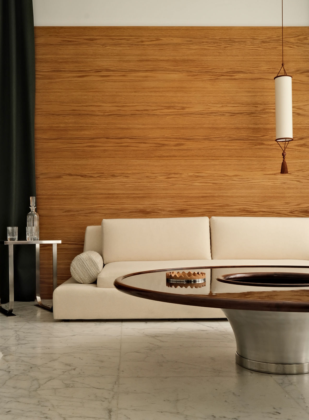 A contemporary living room features a cream sofa set against a wooden accent wall, complemented by the "Loulou" Coffee Table by HAUVETTE & MADANI, which boasts a sophisticated design with lacquered wood and an embossed aluminum base. A side table adorned with glassware sits nearby, while from above, a pendant light softly illuminates the space and partially visible dark curtains add to the atmosphere.