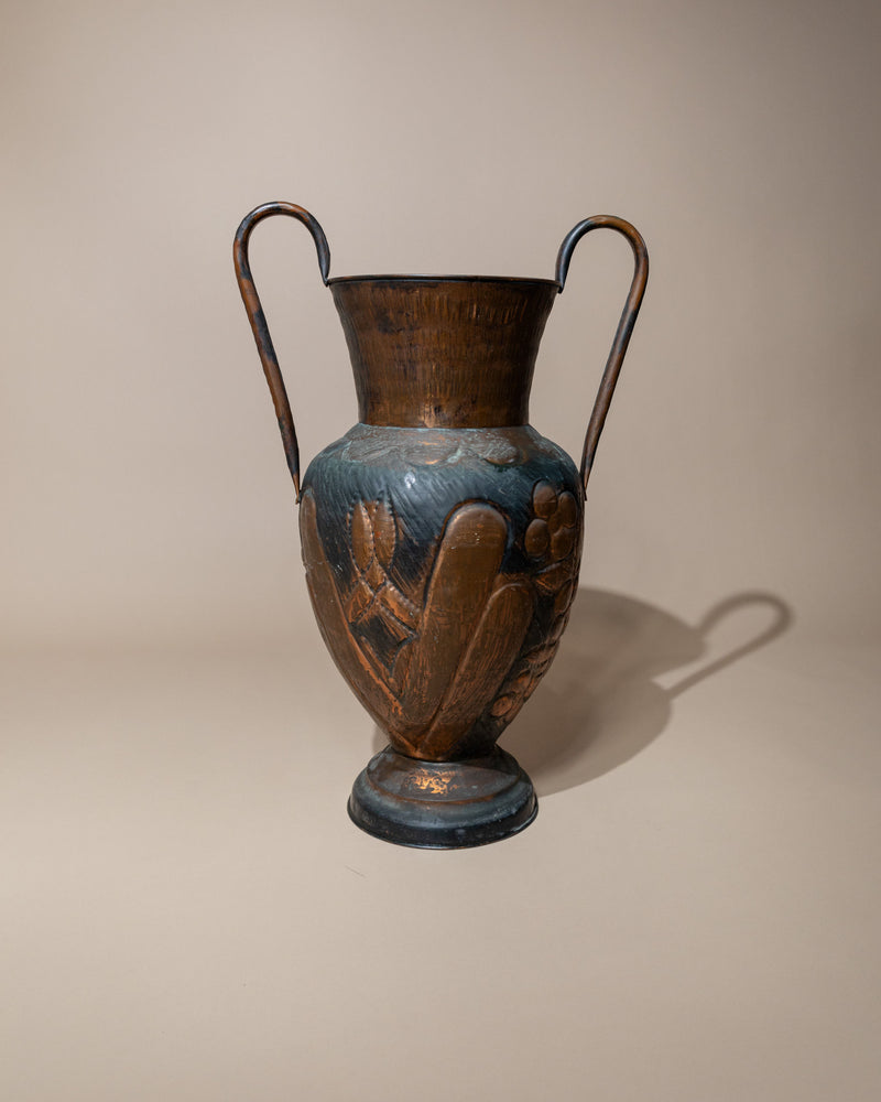 The Copper Vase by Bottega Jacobs, from 1960s Italy, is tall and hand-finished with two curved handles. It features engraved floral patterns and a hammered texture, set against a neutral background and casting a soft shadow to the right.