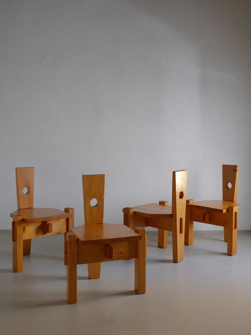 Three 1960s sculptural pine chairs by Erwin Egel from Veter Vintage, each with a tall square back and circular cutout, stand on a light floor against a grayish wall. Their angular design and natural wood finish evoke minimalist, rustic charm.
