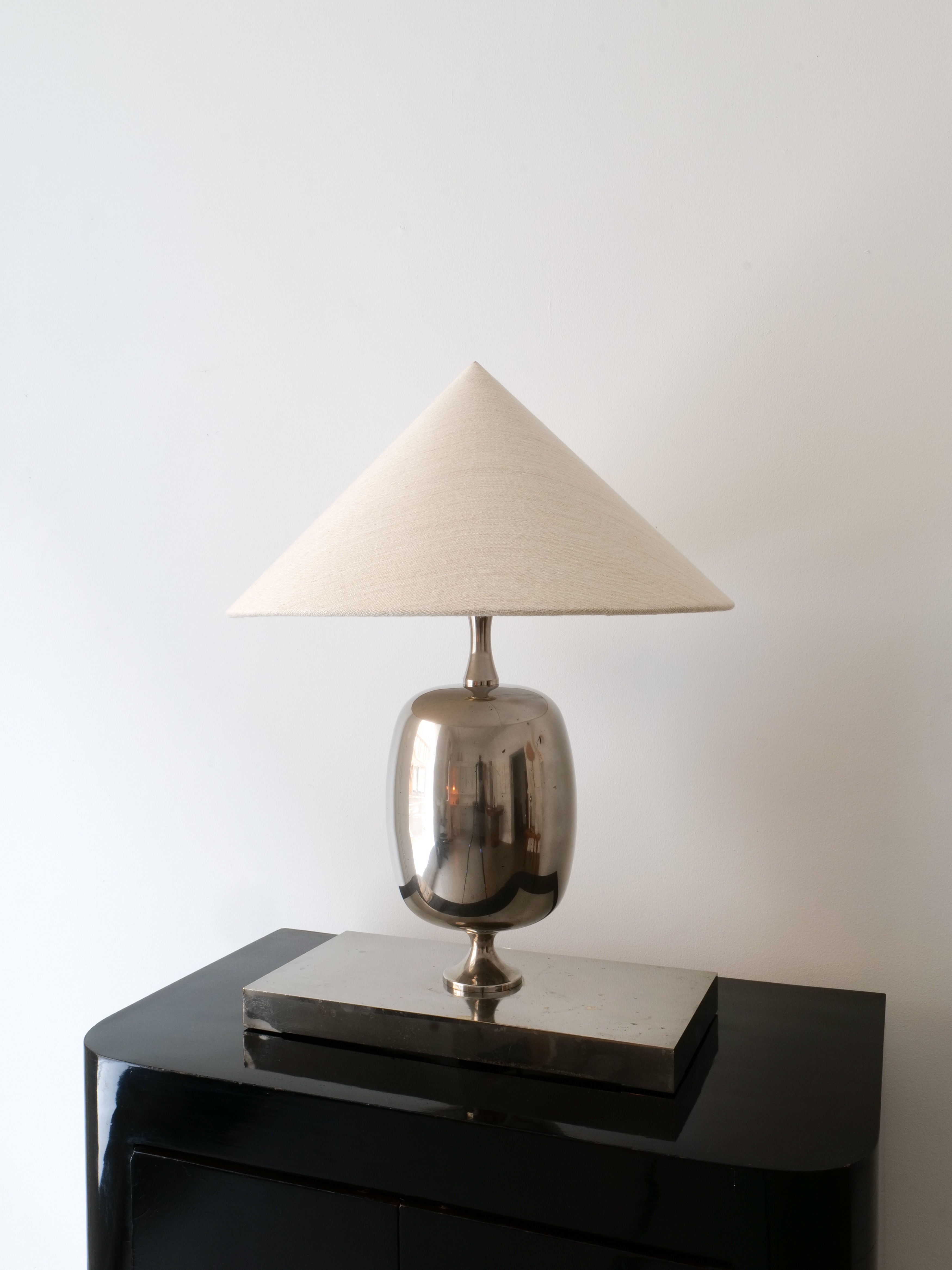 The Barbier Chrome Table Lamp from Collection apart, featuring a chrome-over-brass base and a beige cone-shaped lampshade, rests on a sleek black surface against a plain white wall.