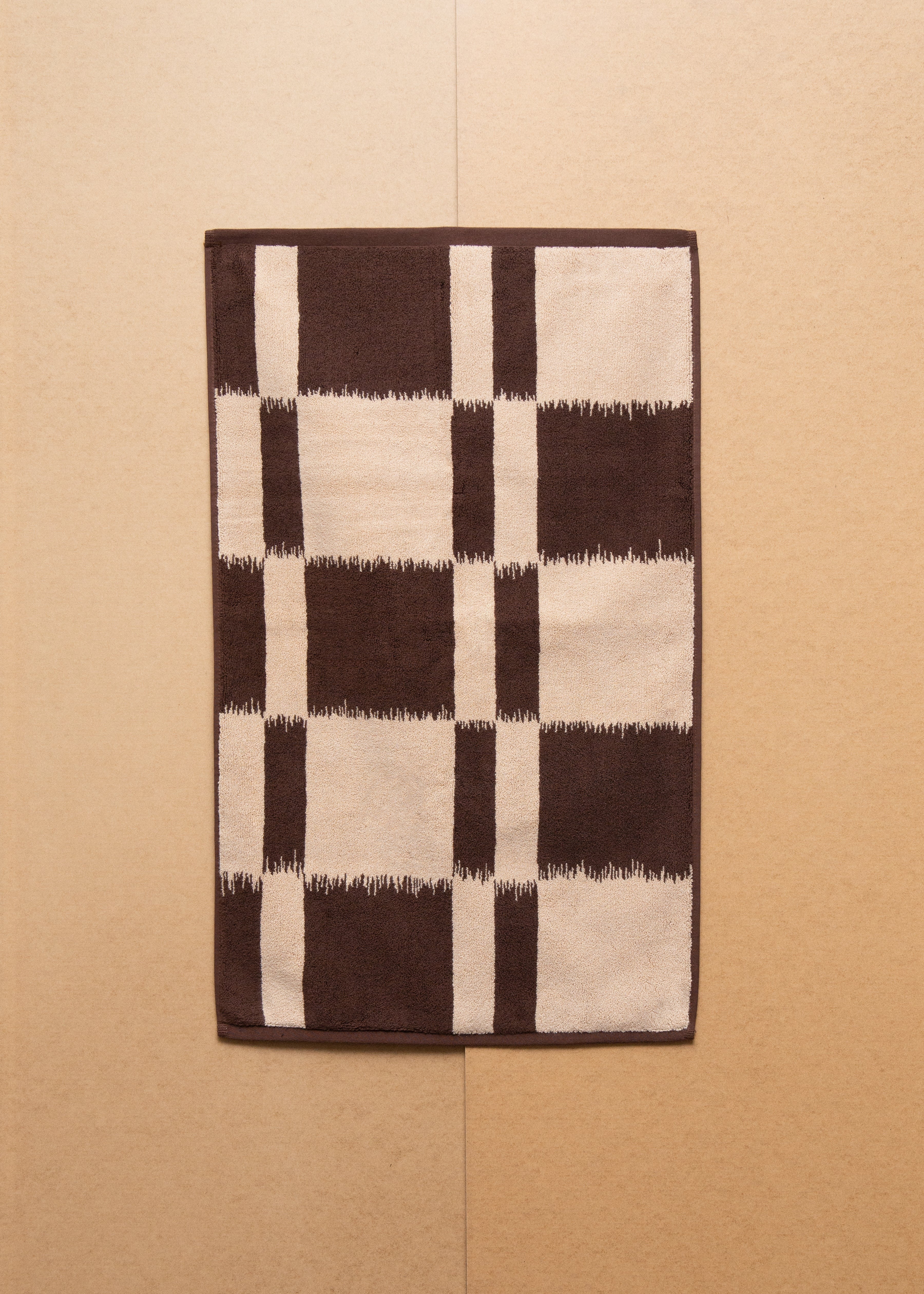 A brown and white Ikat weaving Karin bath mat by Autumn Sonata hanging against a textured beige wall.