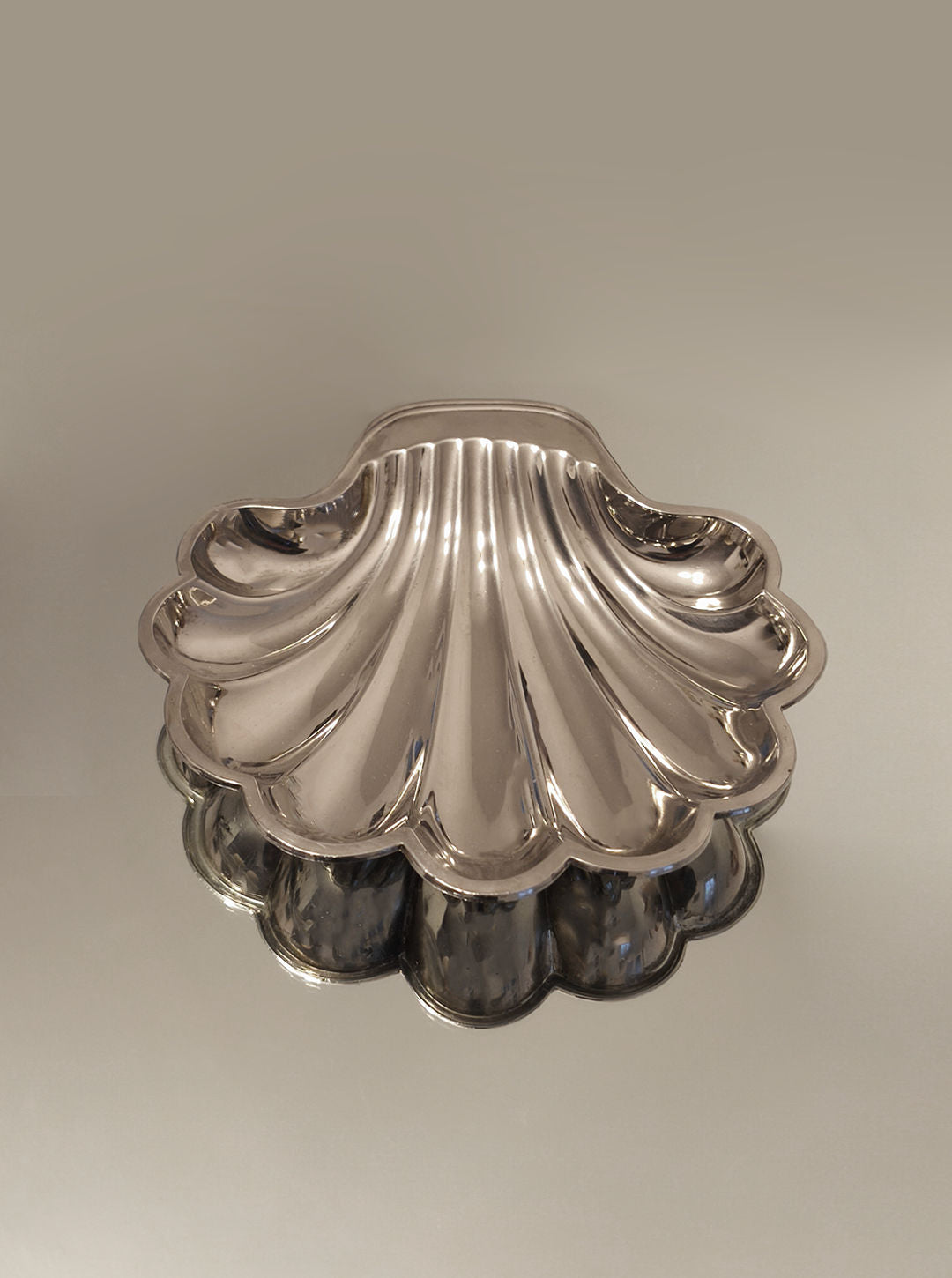 A Shell Shape Vide-poches by Les Objoies, featuring a shiny silver-plated scalloped design, reflects elegantly on a mirrored surface against a soft, neutral background.