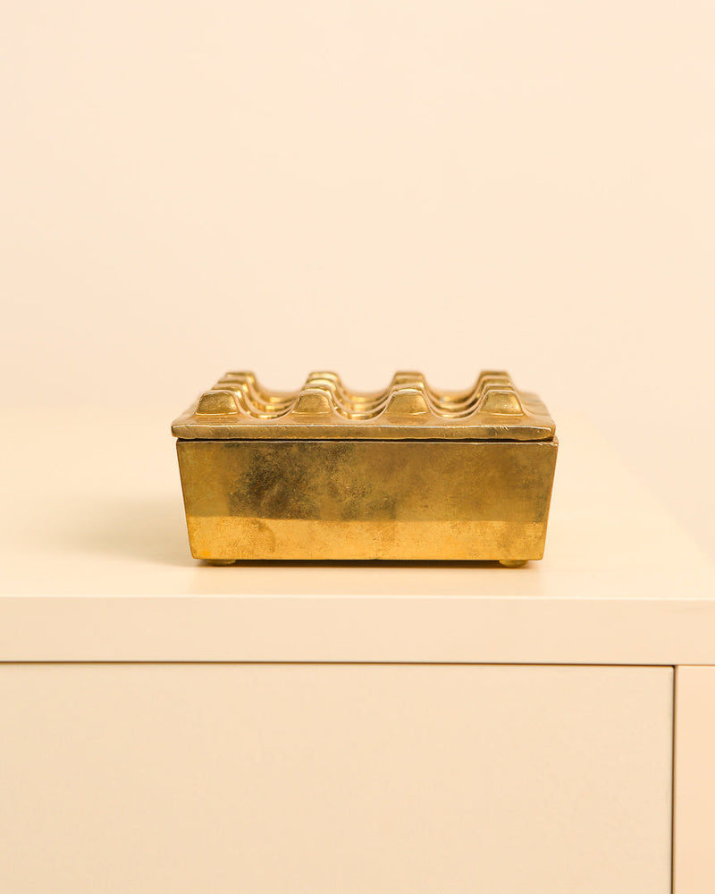 A rectangular, elegant Treaptyque brass ashtray by Holger Bäckström & Bo Ljungberg (70's) sits on a cream cabinet against a pale beige background, featuring a wavy, ridged lid for functional style.