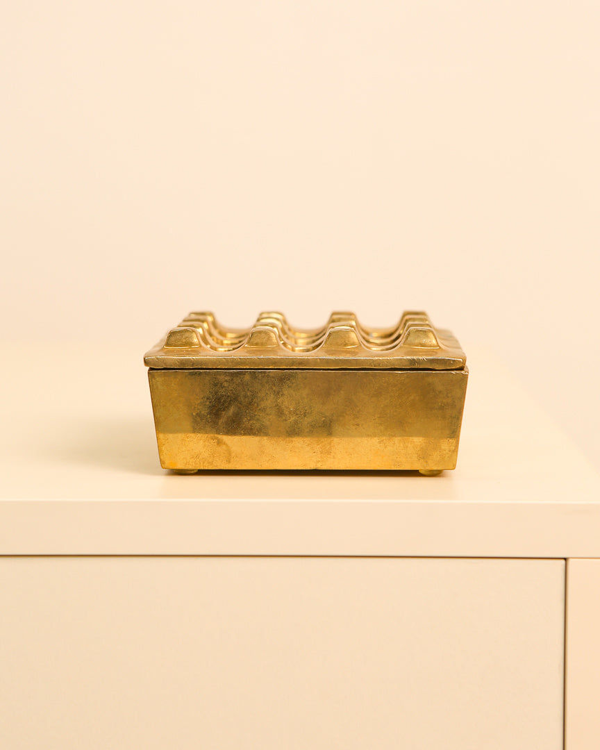 A rectangular, elegant Treaptyque brass ashtray by Holger Bäckström & Bo Ljungberg (70's) sits on a cream cabinet against a pale beige background, featuring a wavy, ridged lid for functional style.