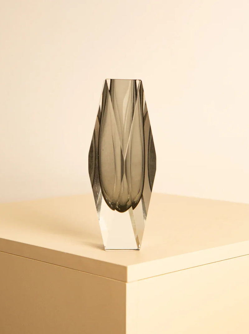 A sleek, geometric glass vase with angular facets, reminiscent of Flavio Poli's Italian glass art, sits on a cream-colored surface against a plain beige background. The Black “Diamond” vase by Flavio Poli for Mandruzzato 70s has a modern, minimalist design and reflects light faintly, showcasing its translucent, smoky gray color.