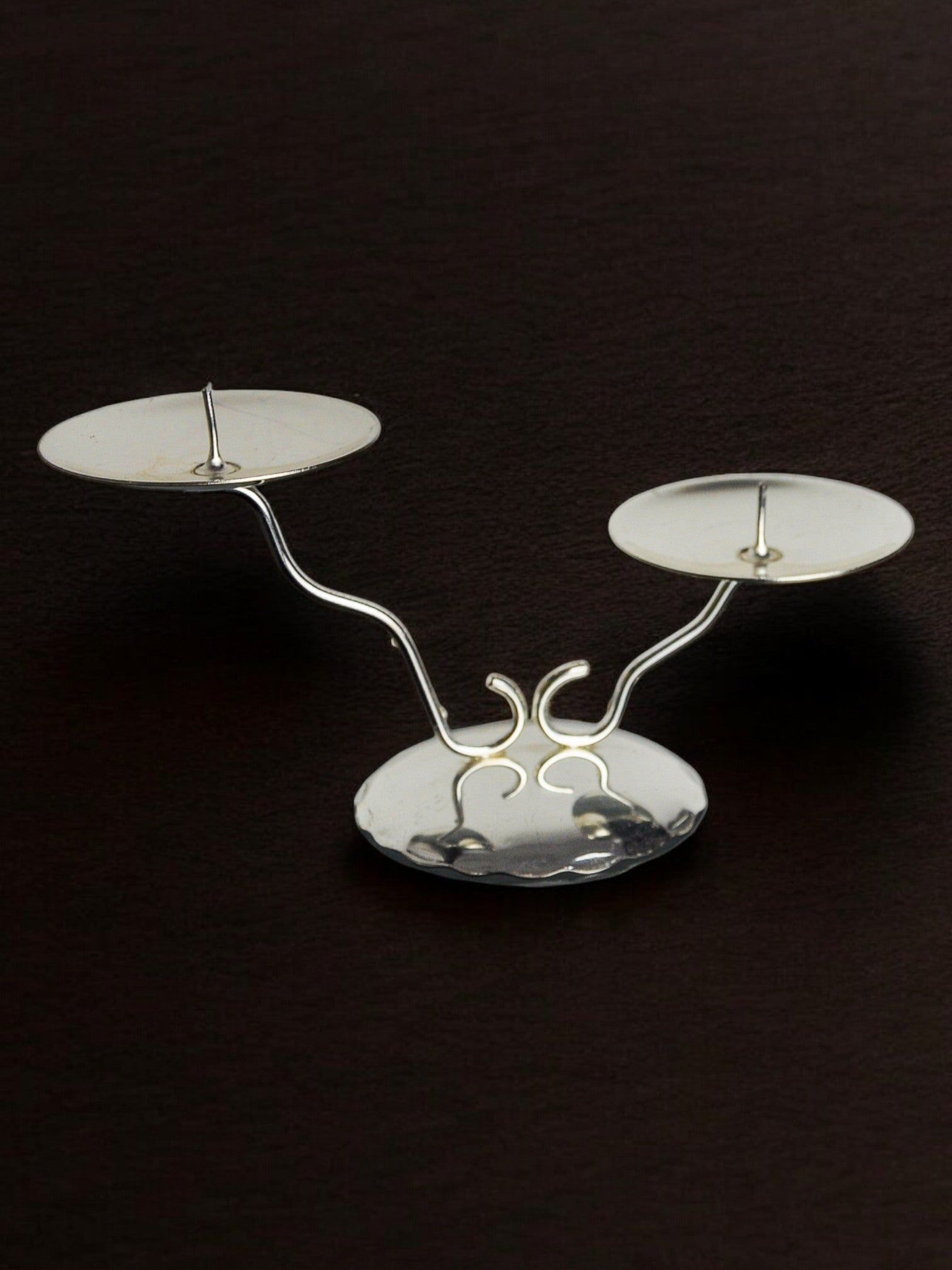 The Art deco 2-flame candlestick by Malamar Studio (Spain, 1980) features a sleek silver structure with two empty round holders on curved arms, one higher than the other, set against a dark backdrop to highlight its asymmetry.