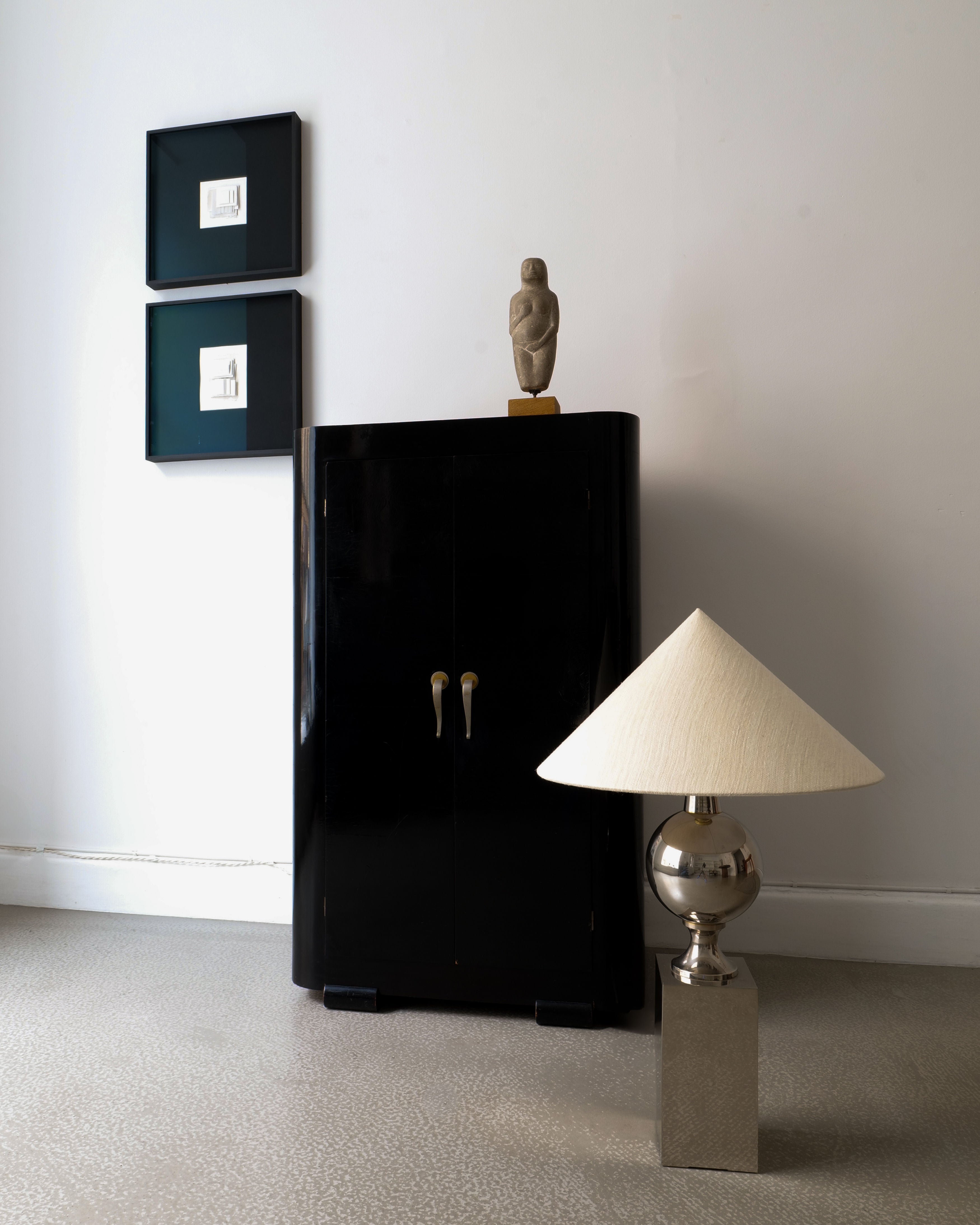 The room showcases a minimalist aesthetic featuring a black cabinet with two doors, upon which a small sculpture is elegantly displayed. Above it, two framed artworks hang with grace. To the right, the Barbier Chrome Table Lamp from Collection apart stands out with its modern design inspired by Philippe Barbier, complete with a cream-colored shade on a square base, all set against a light gray wall.