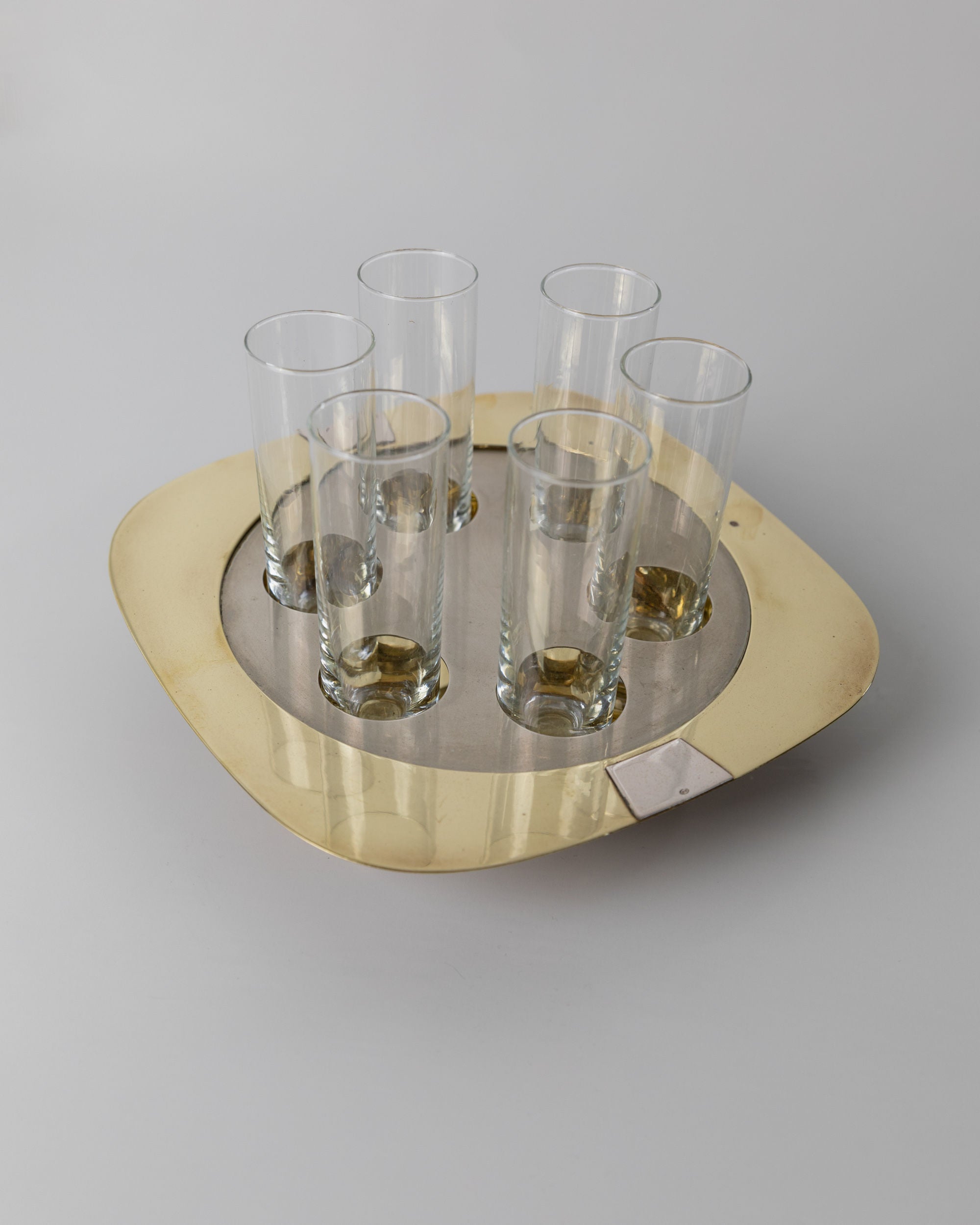 Six transparent shot glasses and a round metallic tray with golden edge from Bottega Jacobs' "Set Shot Glasses and Tray - Mid Century Design 70s" are elegantly displayed on a light gray background, making a striking decorative accent in any setting.