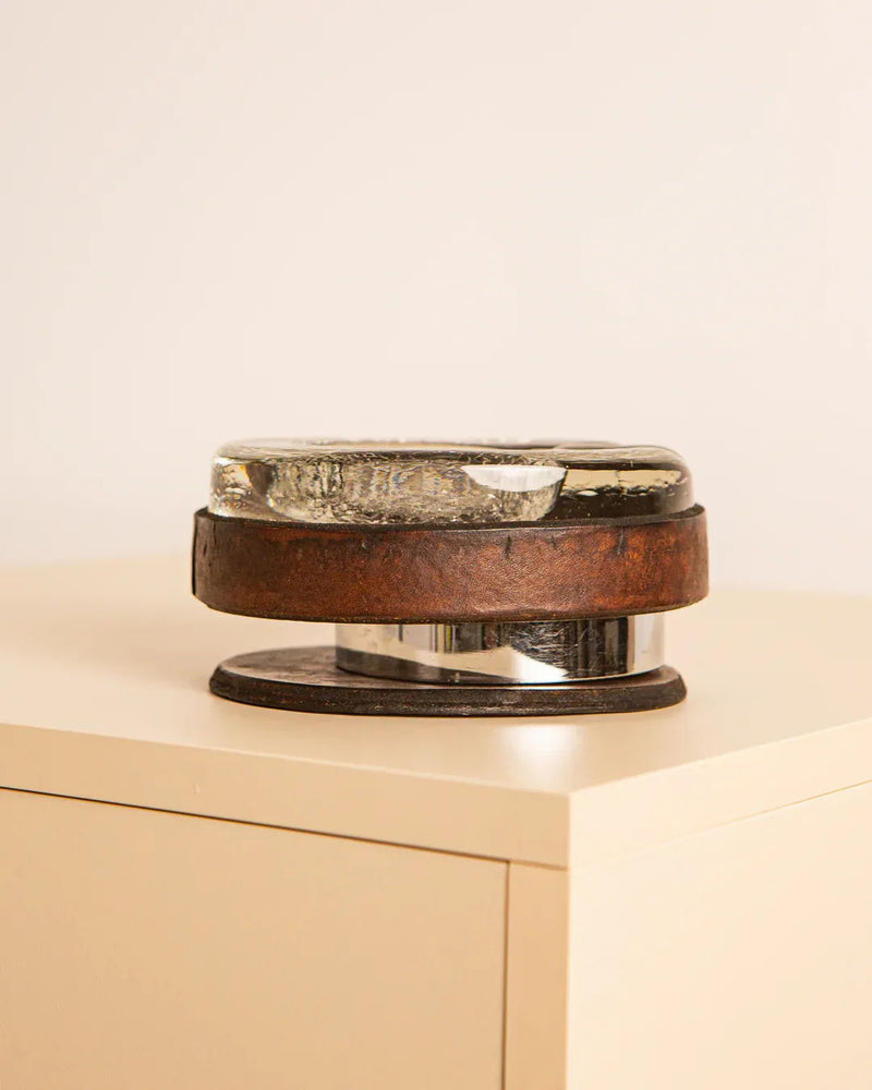 A Treaptyque Murano Glass Ashtray With Leather exudes retro charm, presented on a light wood pedestal. It features a clear, Murano glass oval that evokes an ashtray on a rusty circular metal base, echoing Italian design aesthetics.
