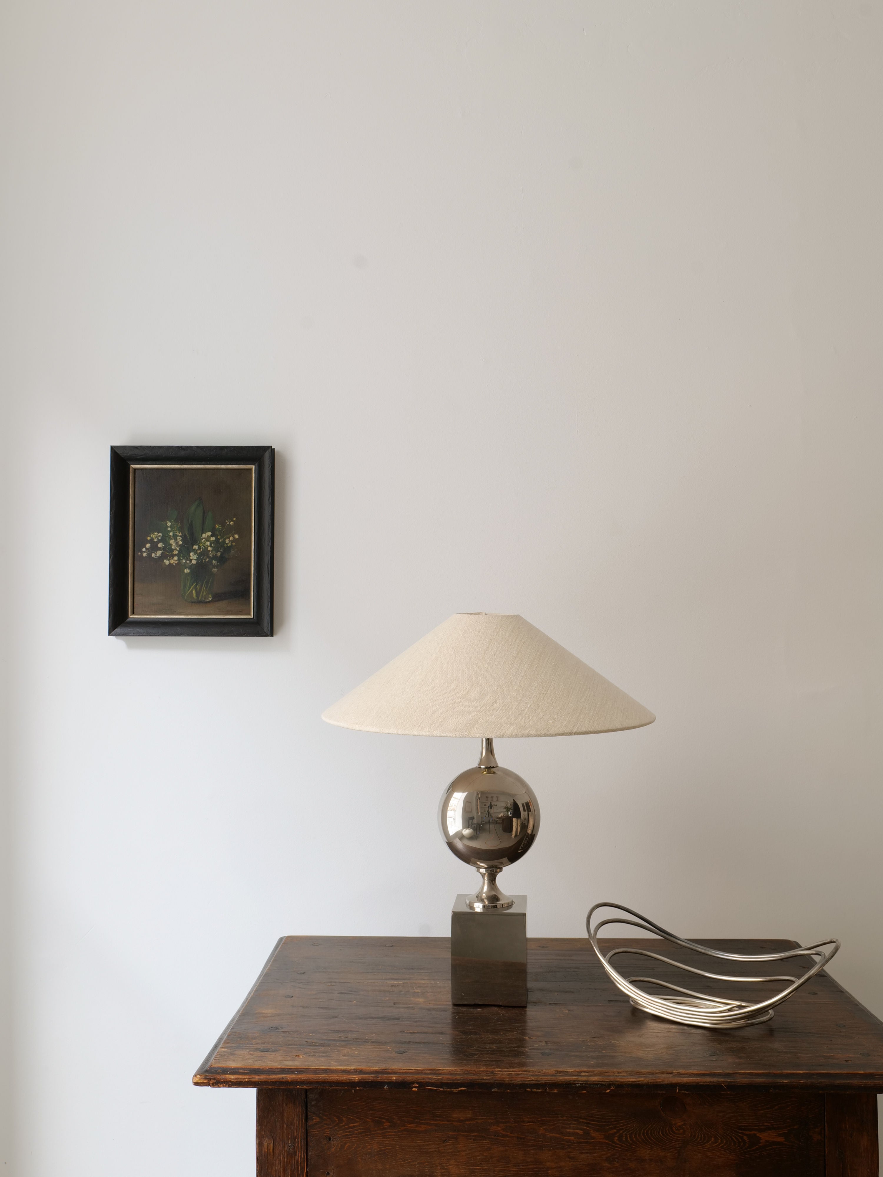 A minimalist room with a framed Lily of the Valley Still Life 1961 by Collection apart hanging on a white wall. Below, a wooden dresser supports a lamp with a polished metallic base and a cream-colored lampshade, alongside a modern, abstract metal bowl.