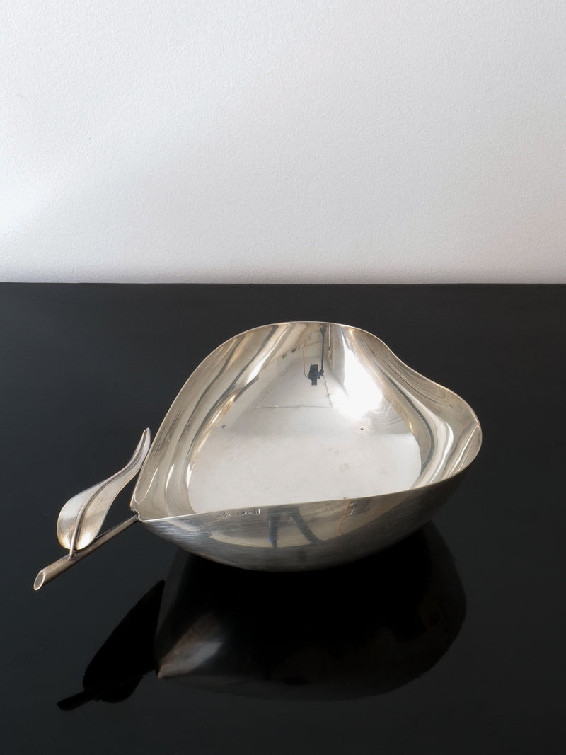 The Christofle Vide Poche Lino Sabattini by Collection apart, known for its collaborations with Christofle, sits gracefully on a glossy black surface. Its asymmetrical design is highlighted by a distinctive wavy handle that resembles a leaf, set against the simplicity of a plain white wall.