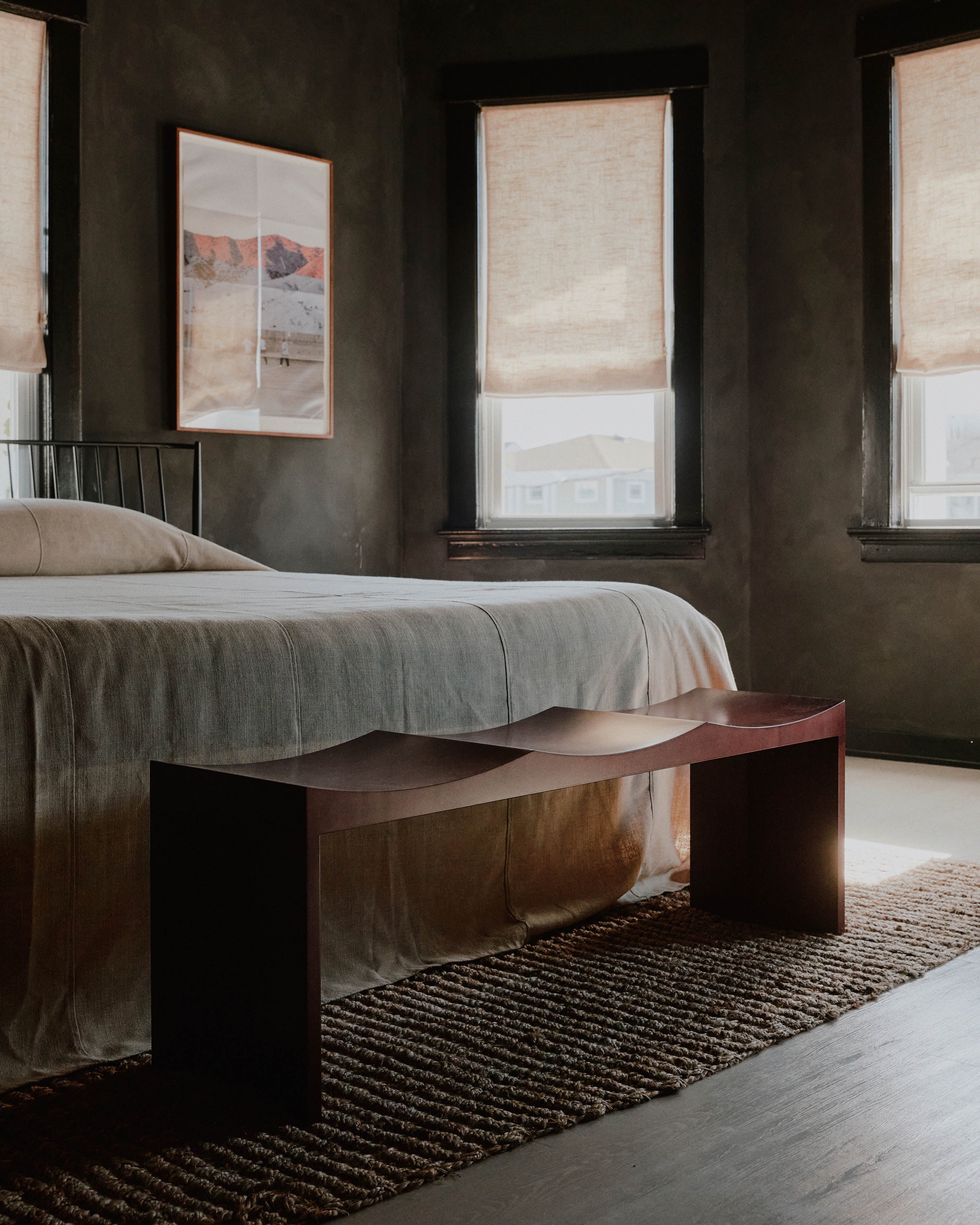 A minimalist bedroom featuring a large bed with neutral-colored bedding, a Juntos Projects Arc Bench at the foot of the bed, and three windows with beige roman shades. The room has dark walls, a striped rug, and a framed landscape picture on the wall, showcasing contemporary furniture handmade in the United States.