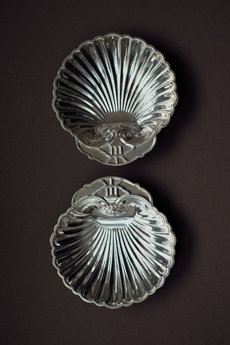 The Set of 2 Silver-plated Shell Trays by septembre studios features ornate, scallop-shaped designs. Displayed vertically against a dark background, they highlight meticulous craftsmanship and add elegance to any dining setting.