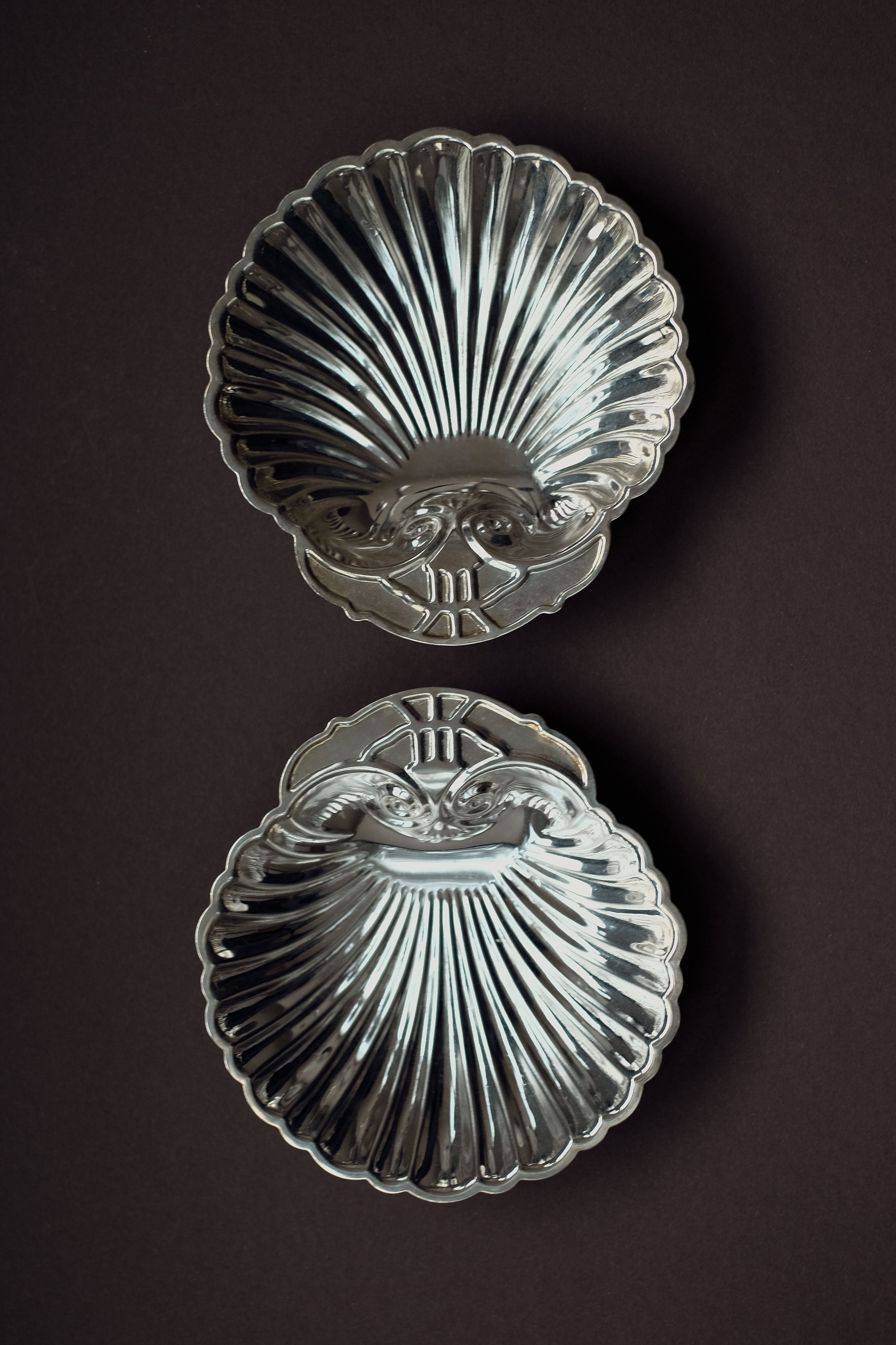 The Set of 2 Silver-plated Shell Trays by septembre studios features ornate, scallop-shaped designs. Displayed vertically against a dark background, they highlight meticulous craftsmanship and add elegance to any dining setting.