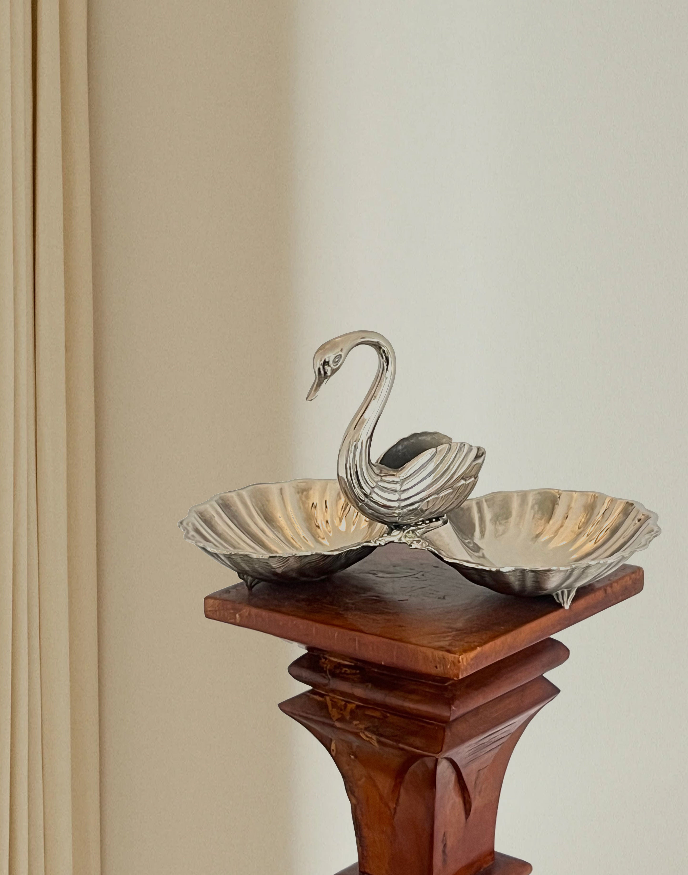 The Porter Studio Ornate Swan Shell Dish, silver-plated with intricate scallop shells, is elegantly showcased on a wooden pedestal against a backdrop of a light-colored curtain and plain wall.