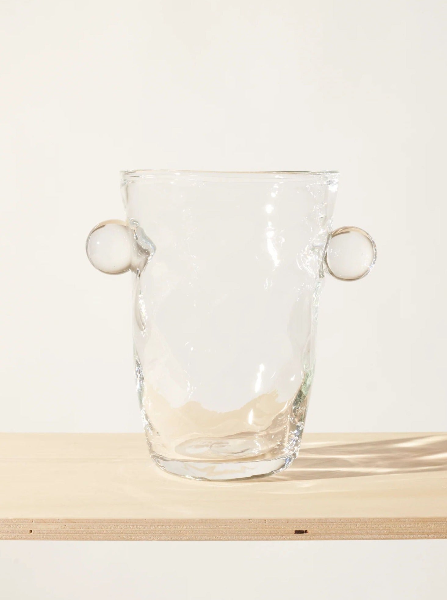 A clear glass Oliver Vase from Akua Objects with a slightly irregular, organic shape stands on a wooden surface. The vase has two circular handles protruding from each side, resembling ears. The overall design is minimalist and modern, showcasing the Bohemian Glassware tradition with subtle reflections and shadows visible on its surface.