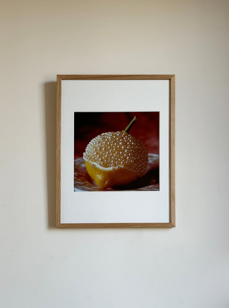 A framed photograph hanging on a plain wall, depicting a close-up of a half lemon covered in clear, bubble-like spheres. The Lemon and Pearls by Adeline Mai combination is set against a dark, textured background. The wooden frame surrounds a white mat with the mixed media photography centered perfectly.
