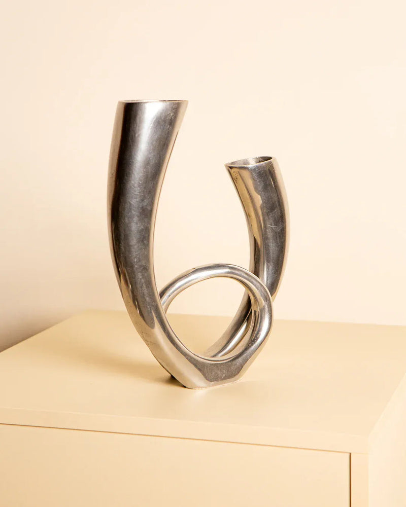 The Treaptyque 80's Italian Aluminum Double Vase features a modern, abstract design with a unique twisting shape. Crafted from cast aluminum, it stands on a beige surface against a light background and features two openings at different heights.