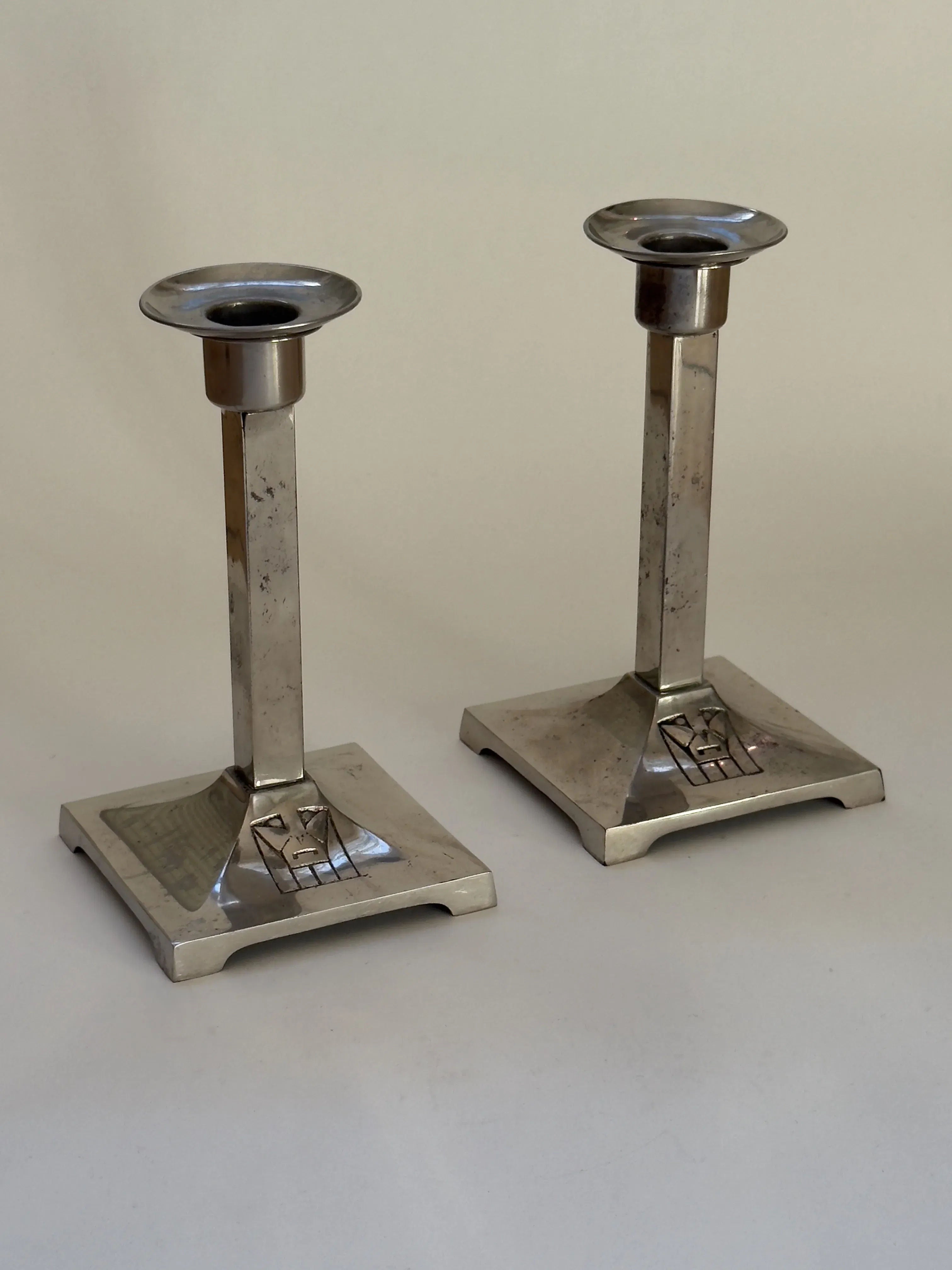 The Pair of Art Deco Candlesticks from Galerie Storm, featuring square bases, tall slender stems, round holders, and an engraved pattern with a slightly tarnished finish, highlights early modernist design with a geometric silhouette from the 20s/30s against a neutral background.