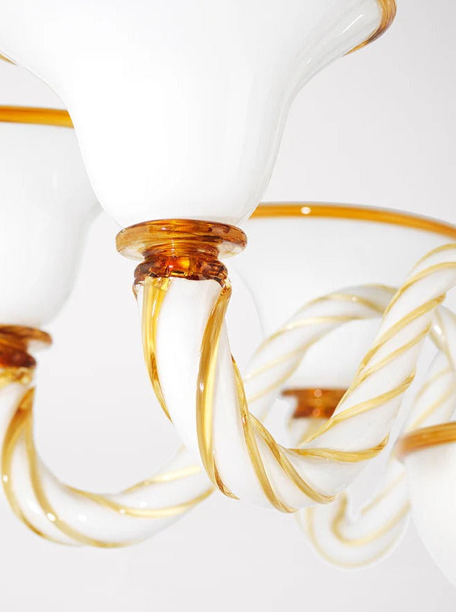 Close-up of a decorative Akua Objects Rosenborg Chandelier with gold-tinted glass accents. The fixture features elegant twisted opaline glass arms and bowl-shaped lamp shades, giving it a sophisticated and ornate appearance against a plain background.