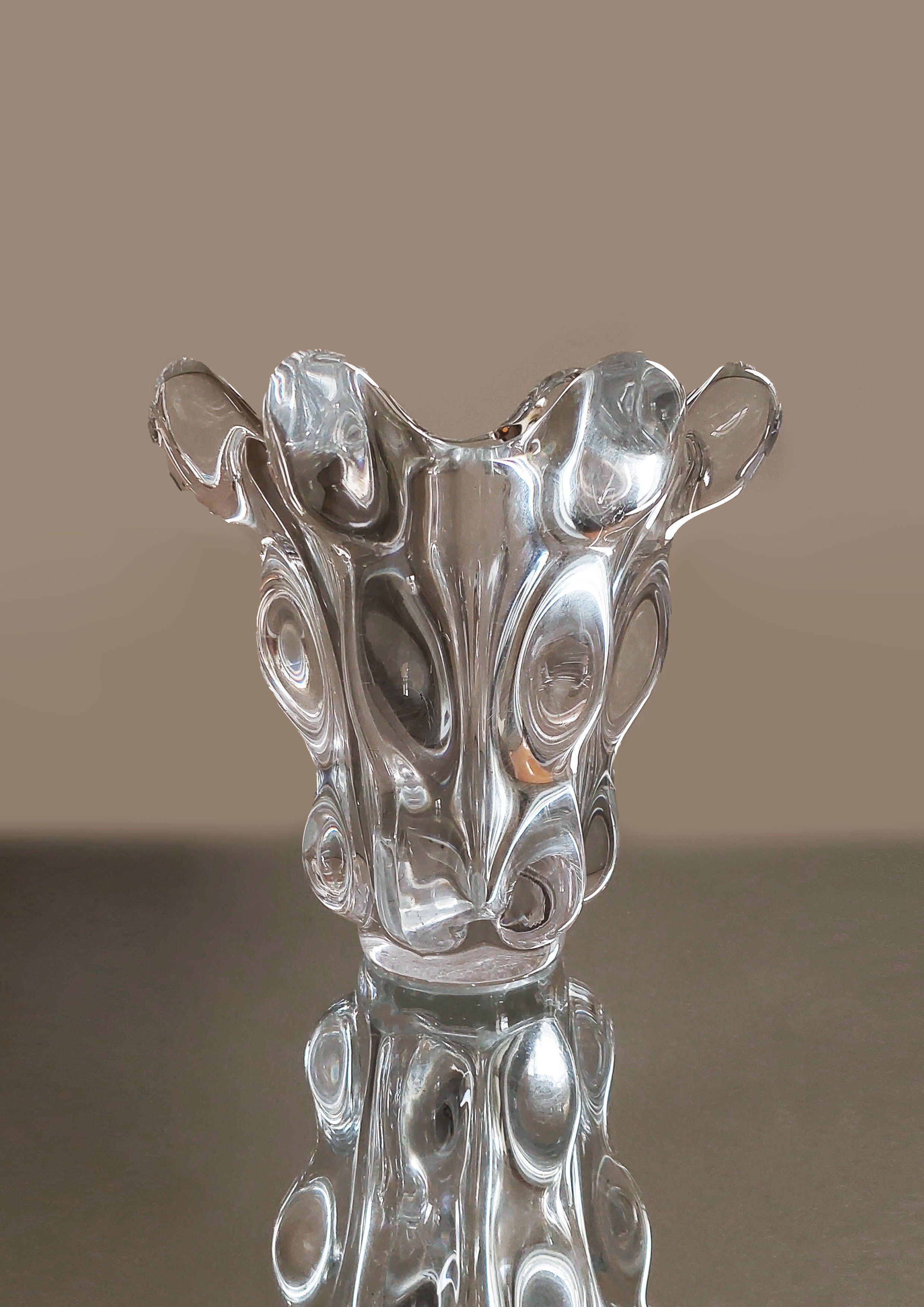 The Les Objoies Crystal Vase, featuring an abstract design with a wavy, circular pattern and an irregular shape, serves as a modern décor piece on a reflective surface against a neutral background.