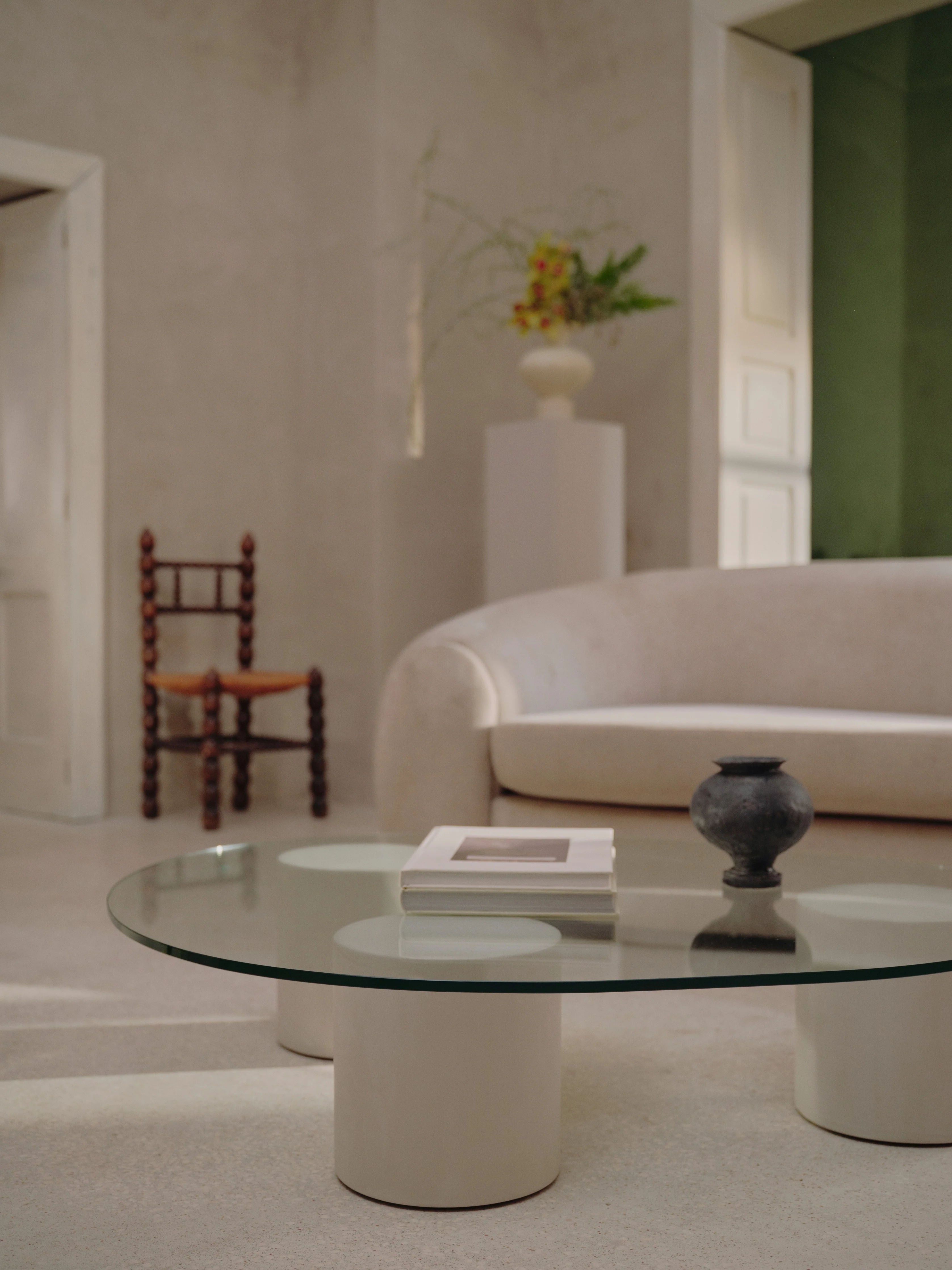 A minimalist living room with a white curved sofa and the "Mattia" Glass Coffee Table from MarlotBaus, featuring a black vase and a book. The room has neutral walls, a wooden chair with a woven seat, and a tall pedestal adorned with flowers.