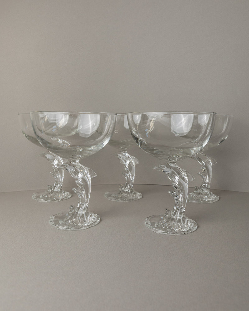 A set of 5 clear Luminarc Delfin champagne coupes by Bottega Jacobs, featuring elegant dolphin-shaped stems, styled against a neutral background. Origin: France, 80s.