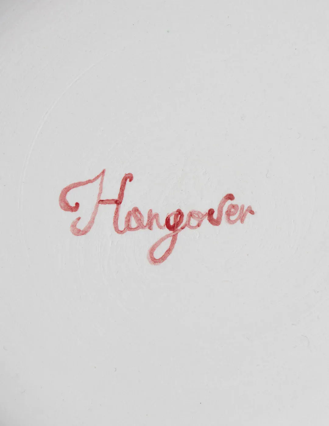 The image features the Musae Studio "Hangover" Fil Rouge Plate, showcasing the word "Hangover" elegantly written in a cursive style with pinkish-red ink on a plain white background, evoking the charm of hand-painted plates from Cilento, Italy.