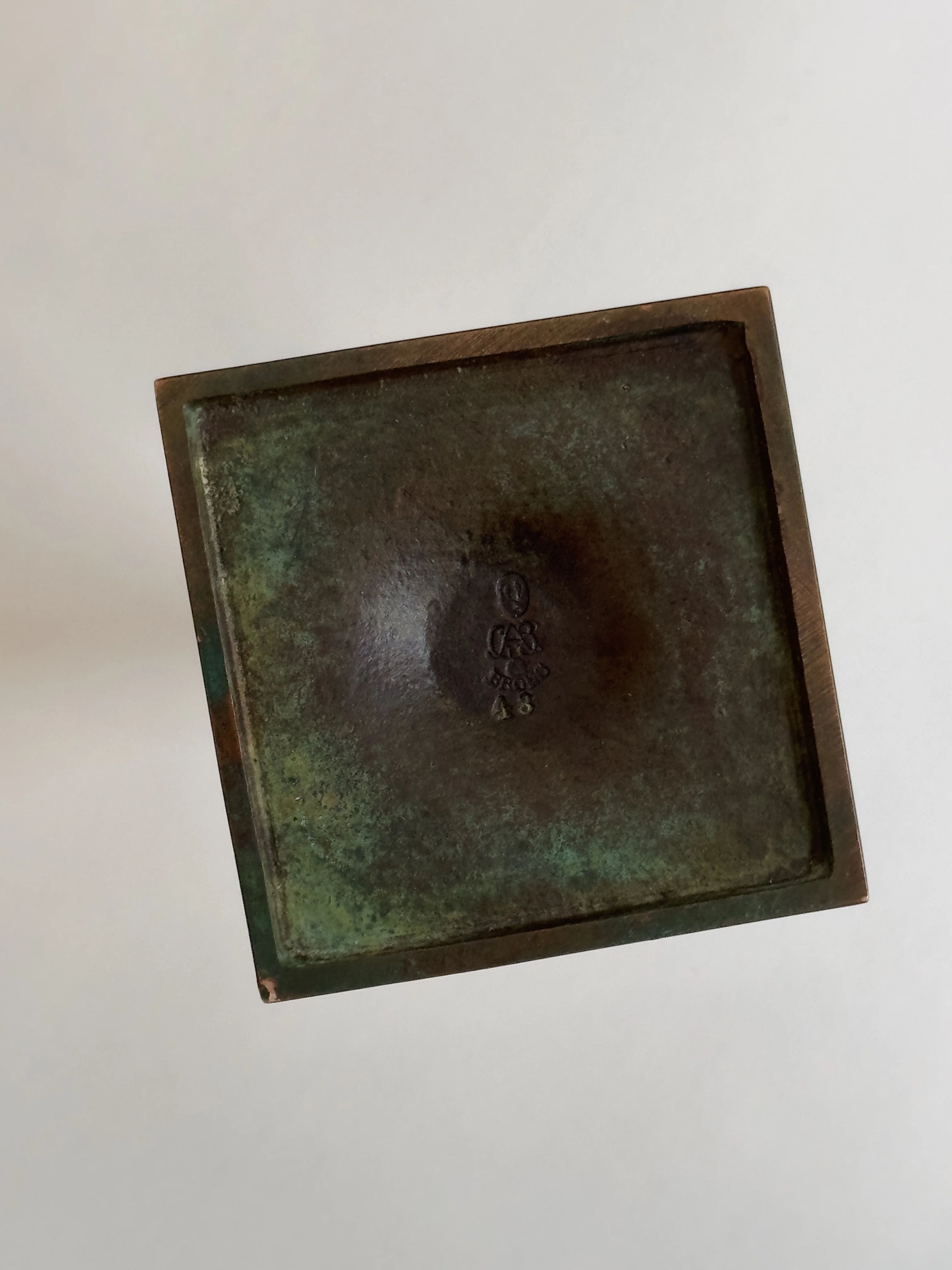 The GAB Bronze vase by Galerie Storm features a square metallic design with a worn green patina, reminiscent of 1930s patinated bronze. Its center displays an embossed symbol with slight fading. The plain, light backdrop enhances its vintage charm and echoes classic GAB artistry.