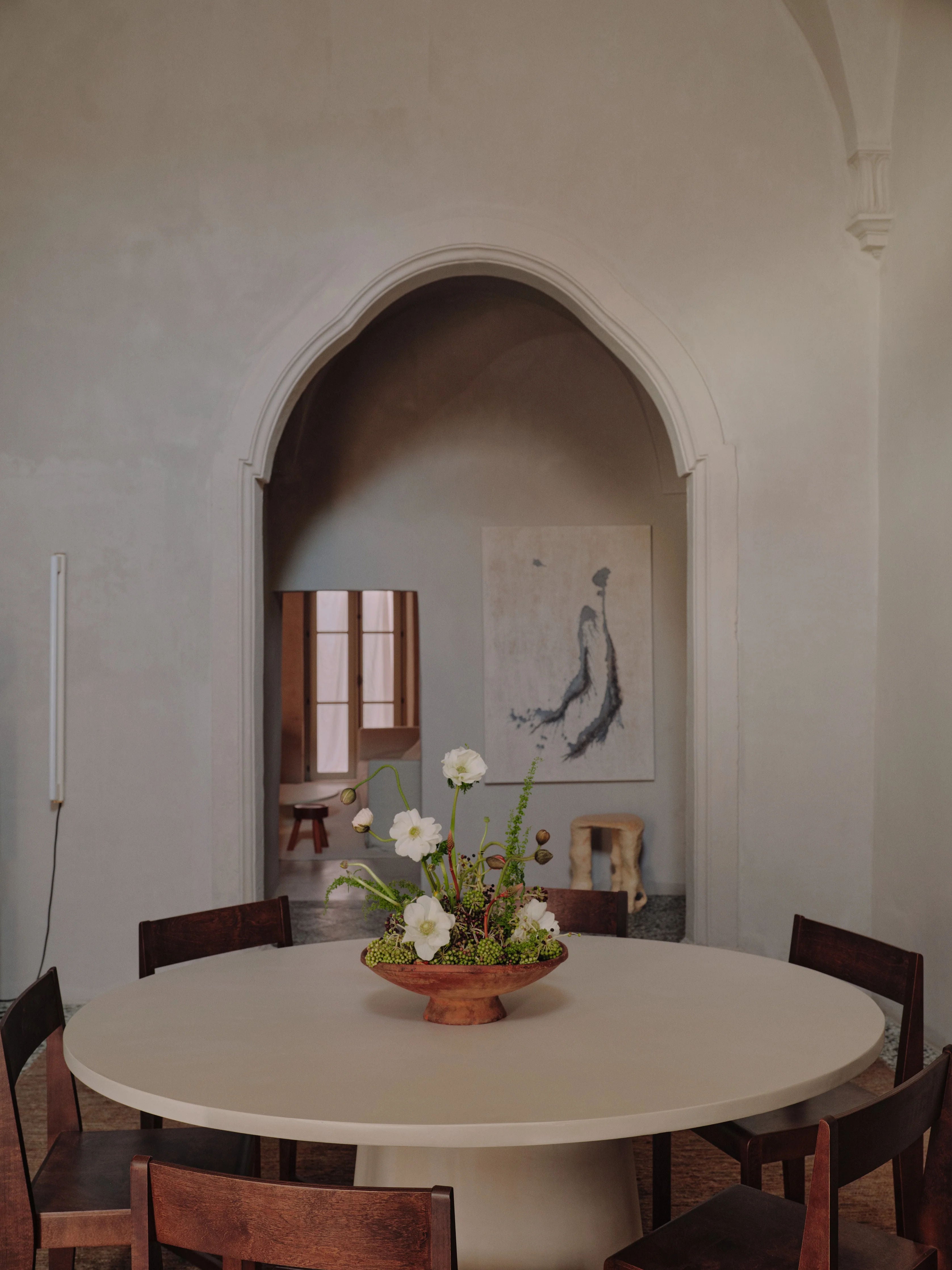 A "Core" Dining Table by MarlotBaus serves as the modern centerpiece in a minimalist dining room, adorned with a floral arrangement and encircled by wooden chairs. An arched doorway opens to a smaller room that features a window and abstract artwork on the wall, enhancing the warm and elegant ambiance.