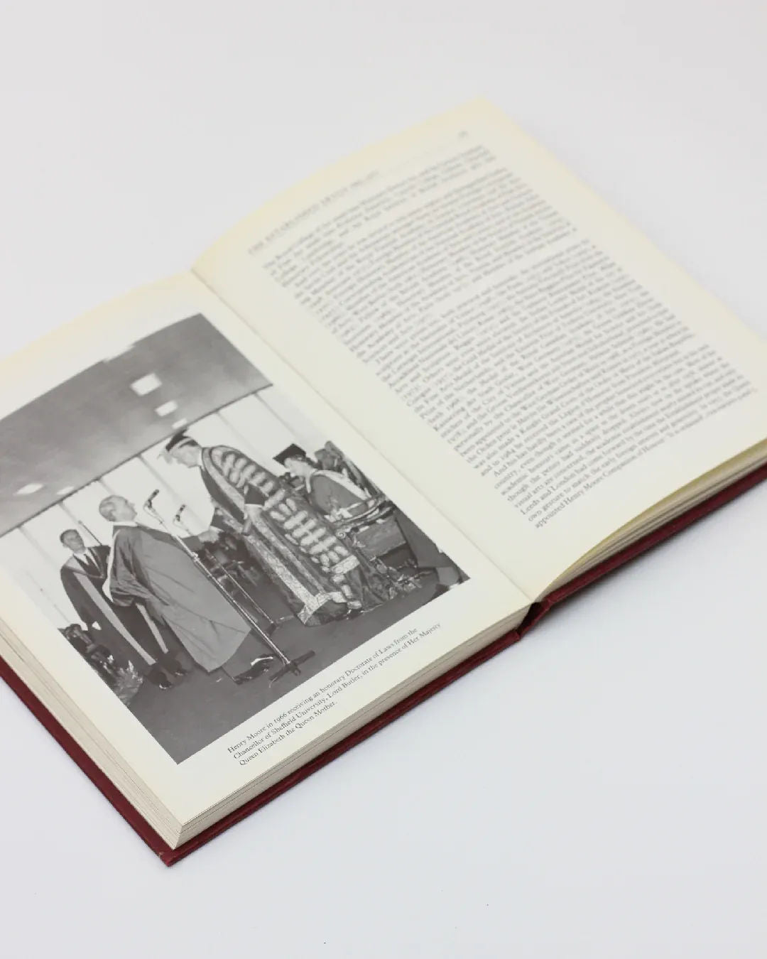 The vintage "Henry Moore, An Illustrated Biography" from Boga Avante Shop is open on a white surface, displaying a black and white photograph of a formal ceremony with people in academic robes on one page, while text occupies the other. The scene evokes memories of scholarly gatherings captured by Henry Moore.