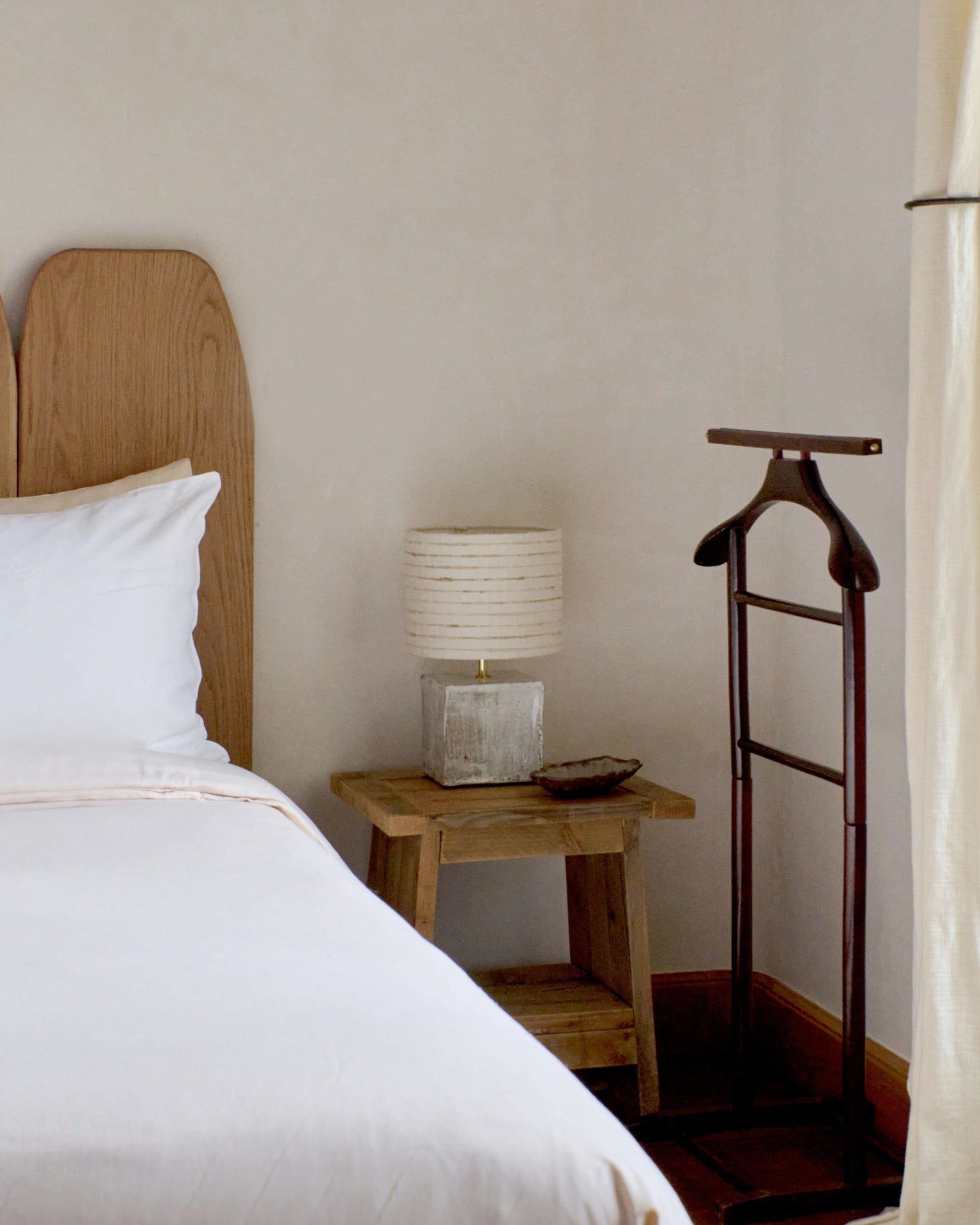 A neatly made bed with a wooden headboard sits beside a small nightstand holding the Project 213A Arouca Table Light Coral White and a dish. By the window with cream curtains, a wooden valet stand stands next to the nightstand.