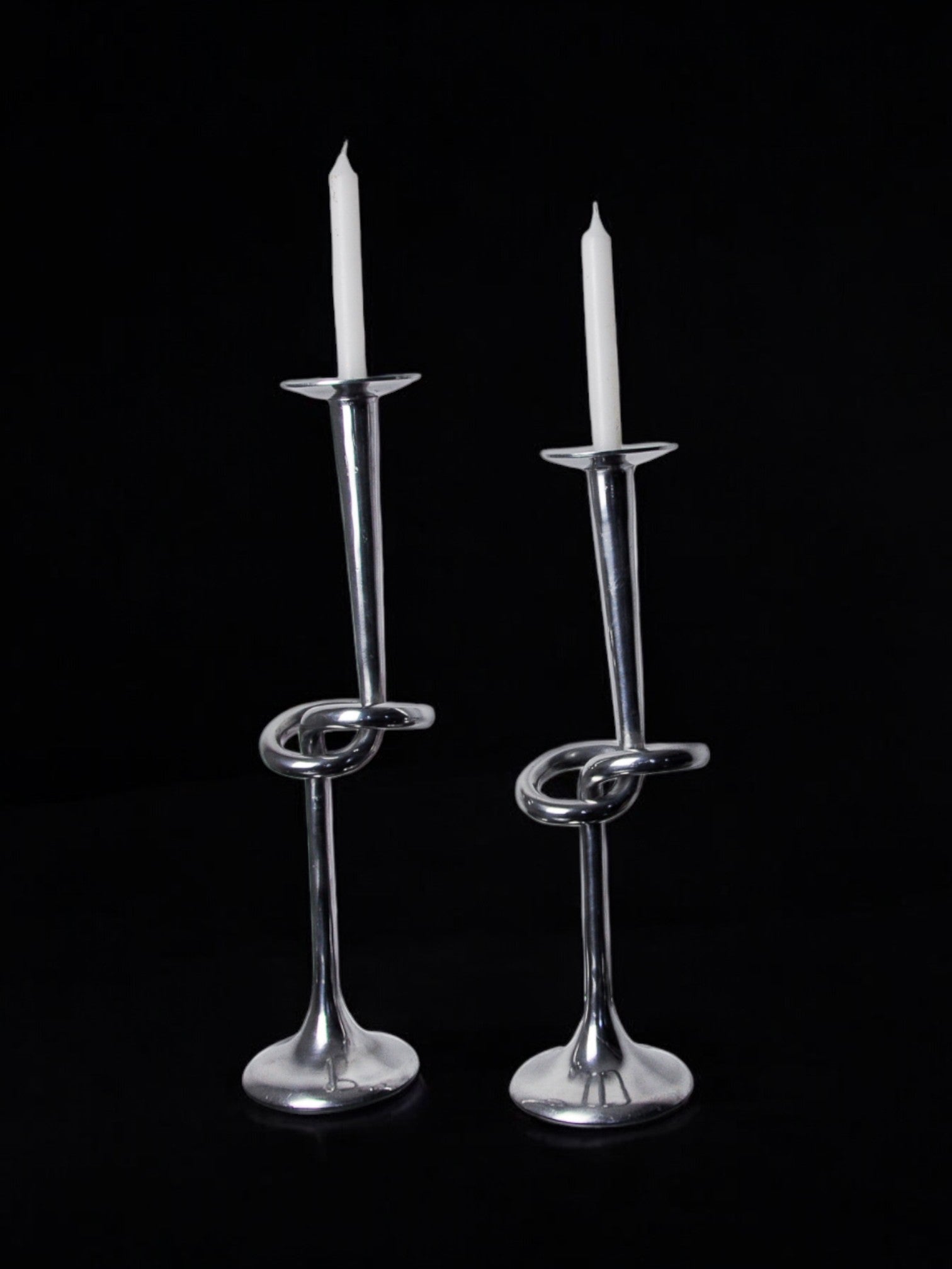 A pair of XL knotted aluminum candlesticks by Malamar Studio, Italy, 1980, each featuring a twisted looped design and holding tall white candles against a black background.