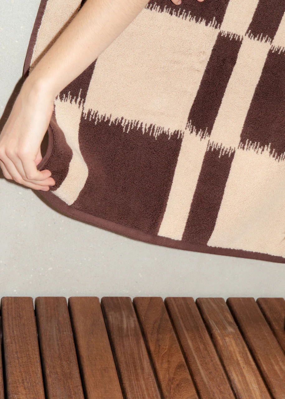 A person's hand pulling up a corner of a brown and beige Ikat weaving rug to reveal a wooden slatted floor underneath the Autumn Sonata Karin Bath Towel.